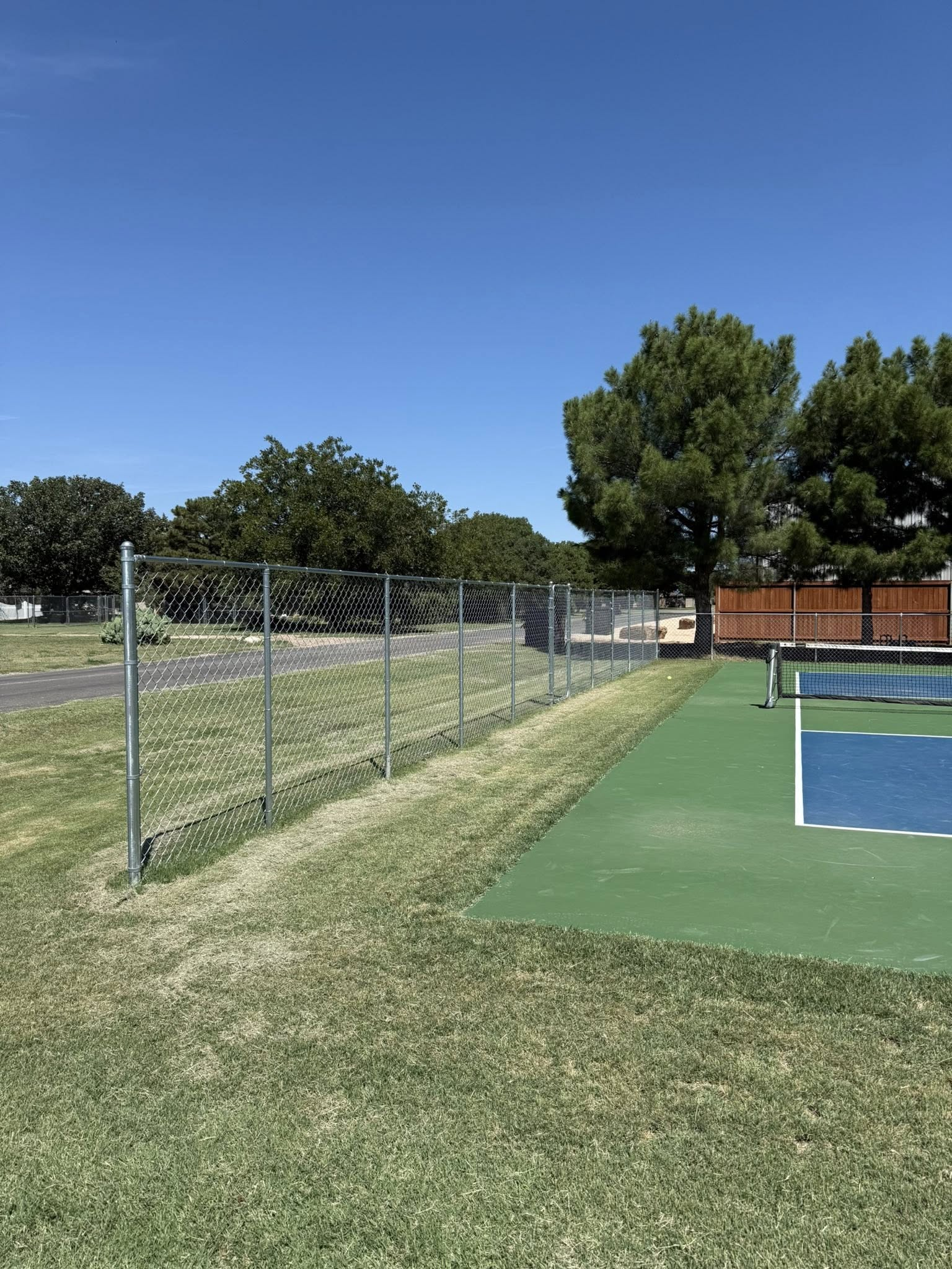 Pickleball court with chain-link fence, green and blue surface, grassy area, trees, and clear blue sky.