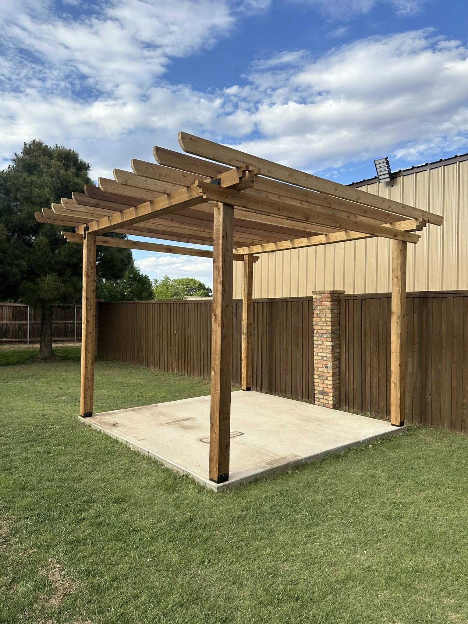 Wooden pergola over a concrete patio in a backyard with green grass, a wooden fence, and a blue sky.