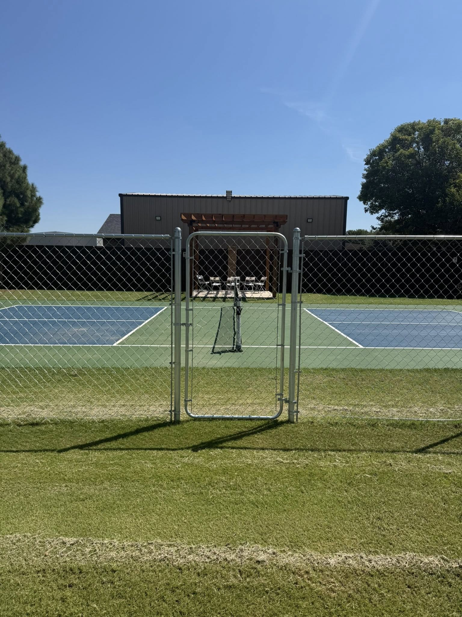 Open gate to a tennis court with green grass, blue court, chain-link fence, and a building in the background.