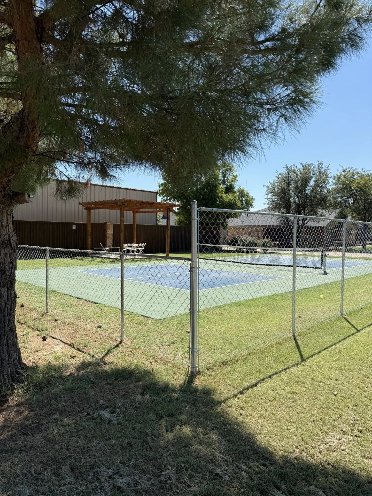 Fenced-in public swimming pool with a shaded pergola and picnic table in the background, under a blue sky.
