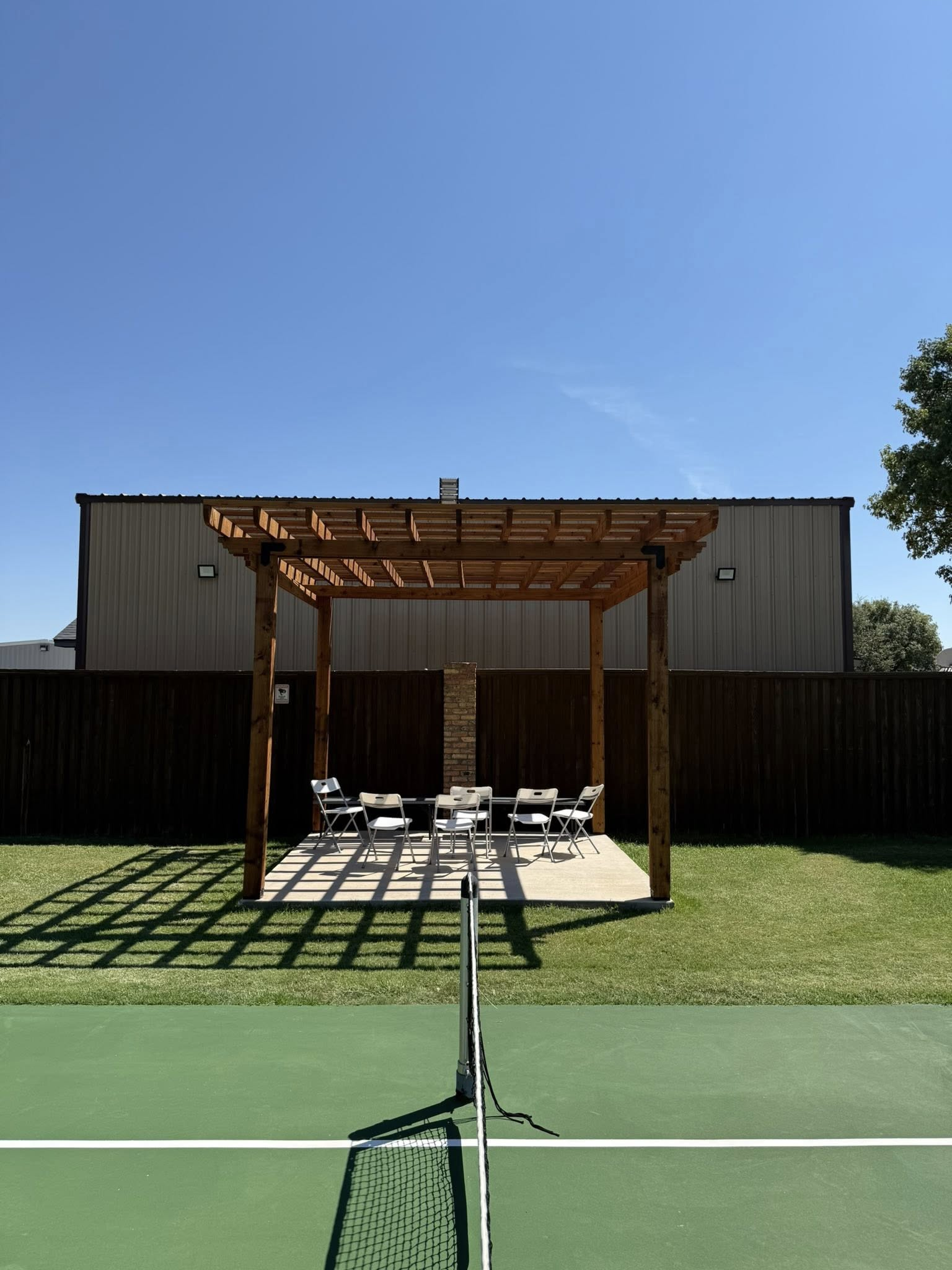 A wooden pergola with seating on a green tennis court, behind a net. Metal building and fence in background.