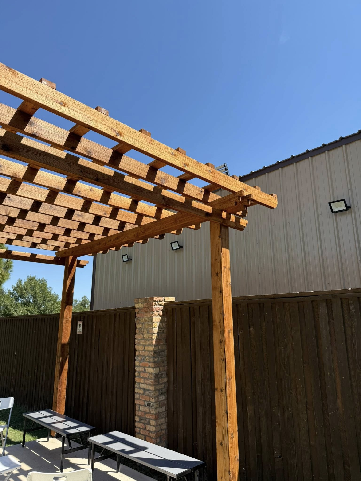 Wooden pergola over tables, with a brick pillar, wooden fence, and building under a blue sky.