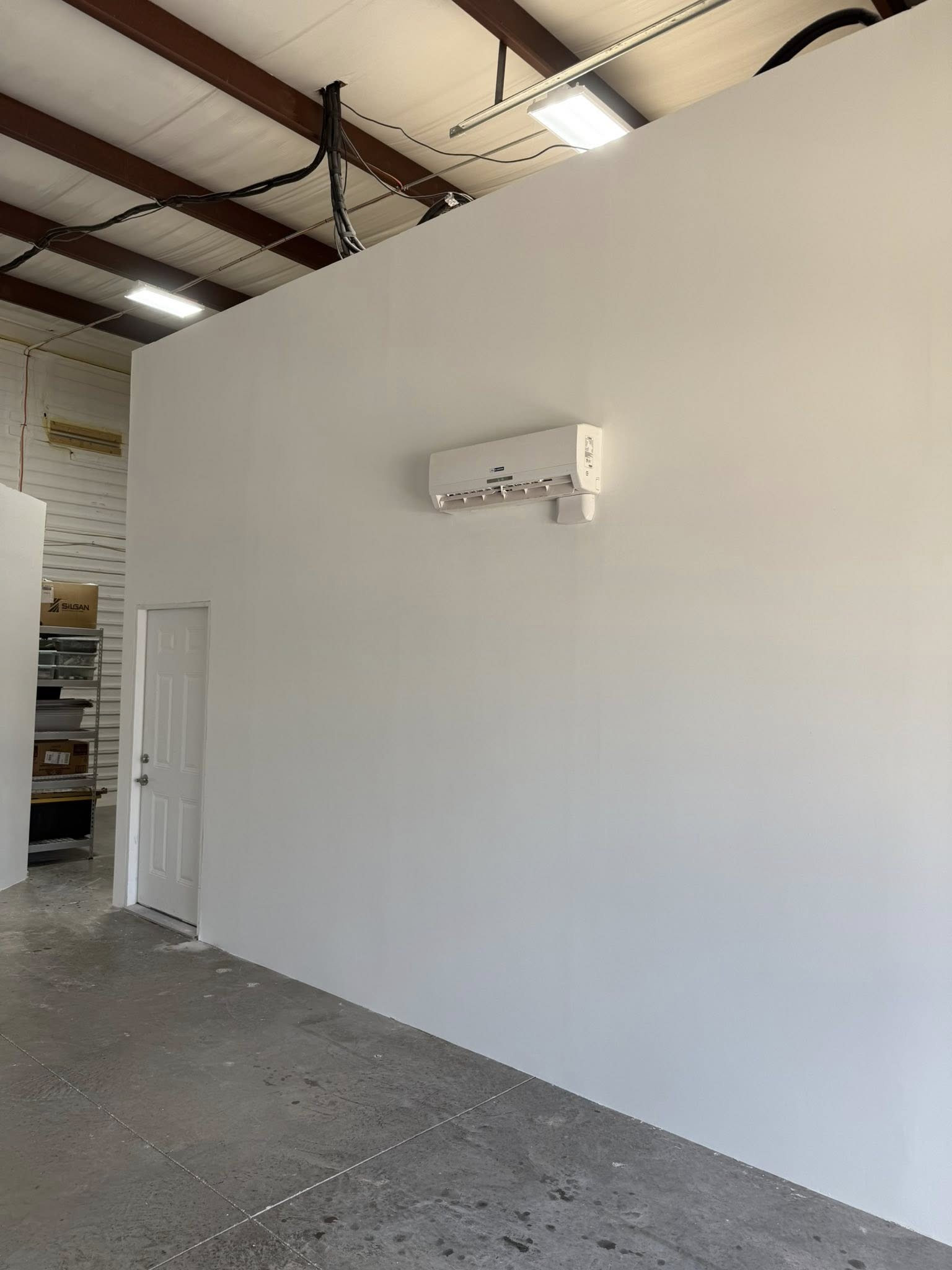 White-walled room with a door and air conditioning unit, possibly a warehouse or workshop space. Concrete floor.