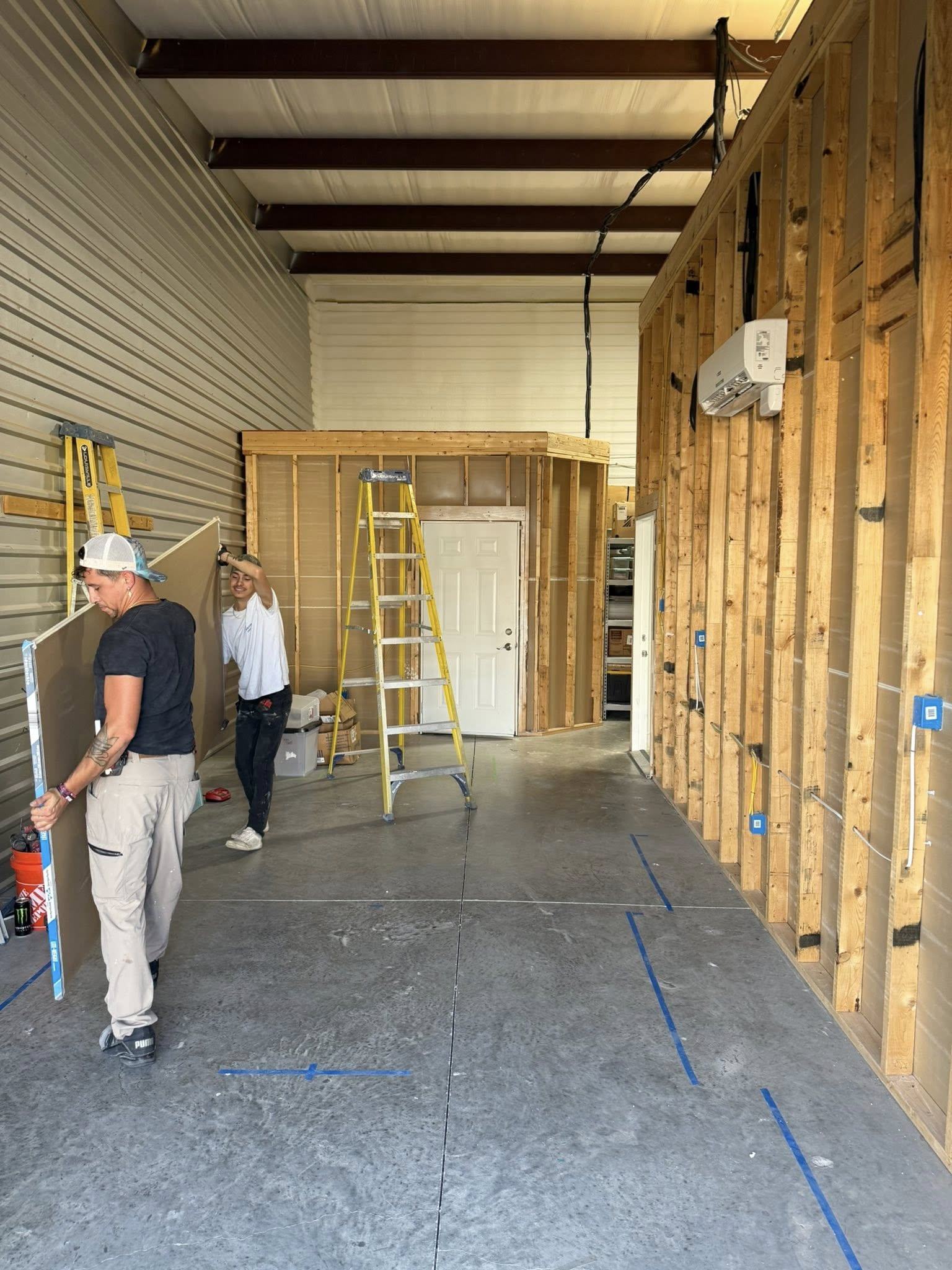 Two people installing drywall inside a warehouse. Framing and studs are visible.