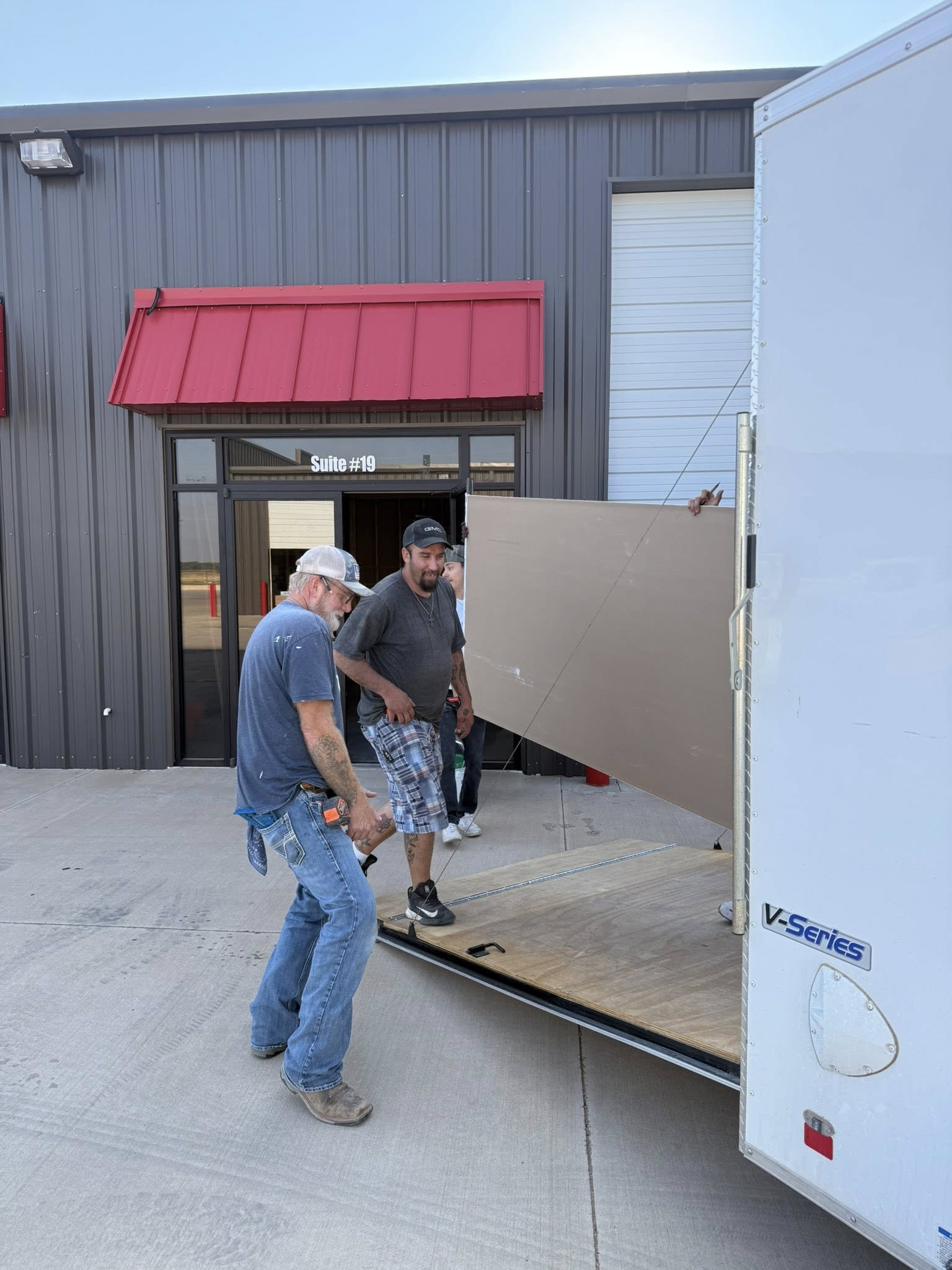 Two men loading a large board into a white trailer outside a building with a red awning.