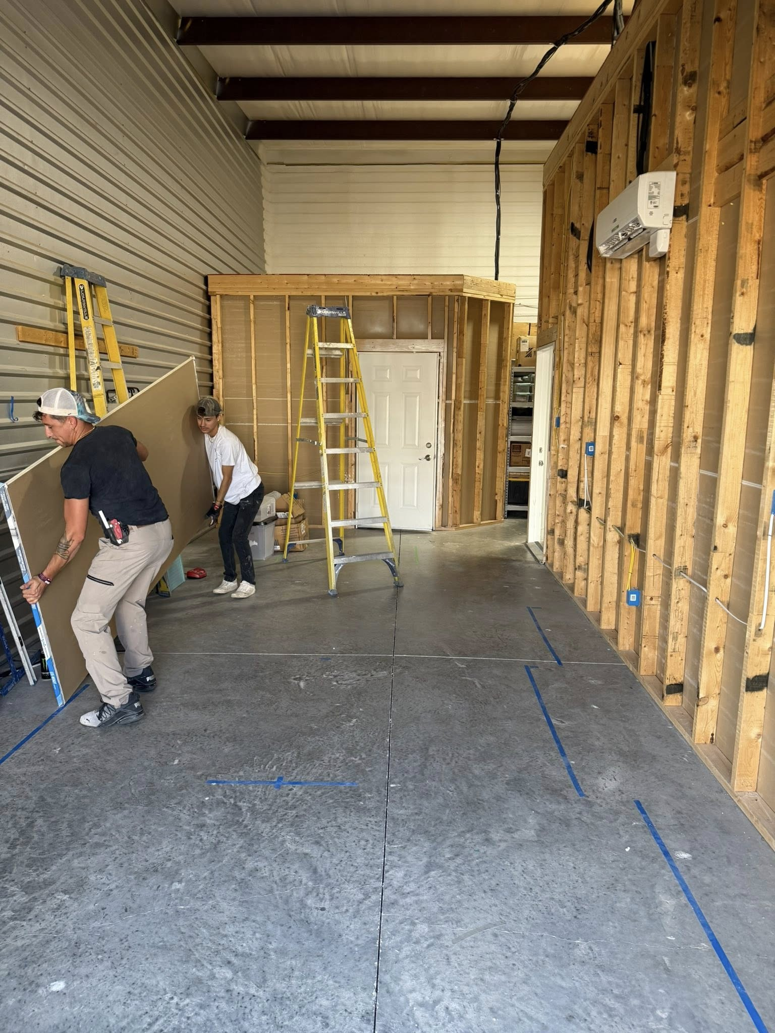 Two people installing drywall in a construction area; wooden frames, ladder, gray floor and walls.