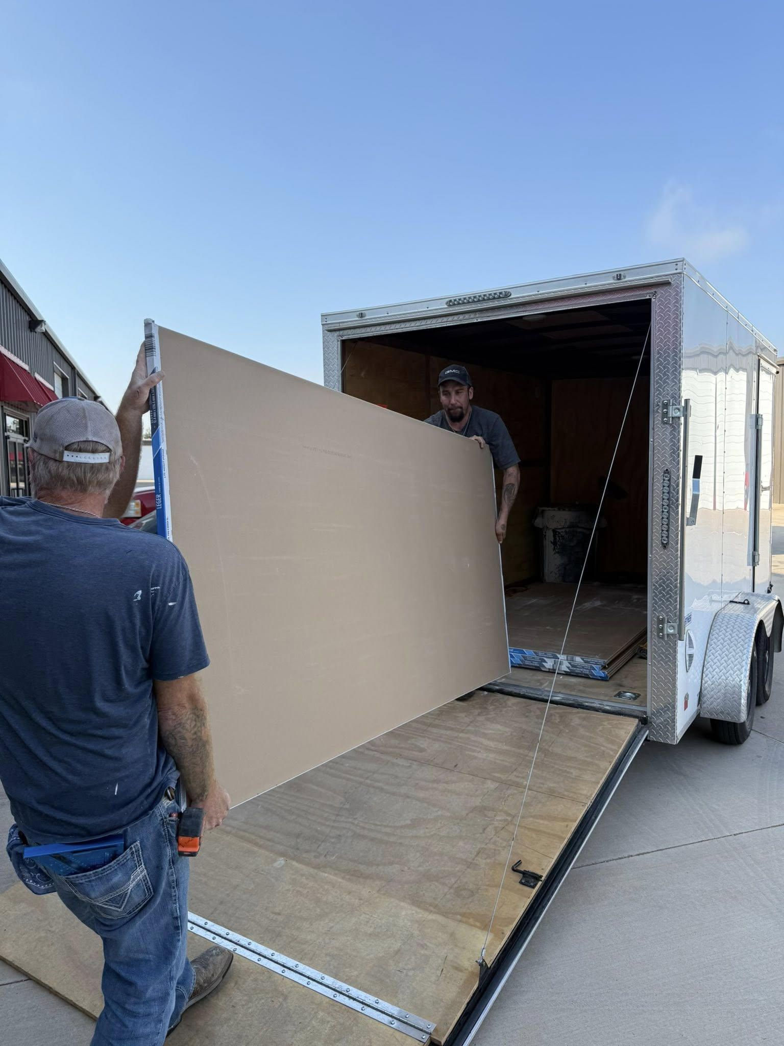 Two men loading large beige drywall sheet into a trailer on a ramp under a blue sky.