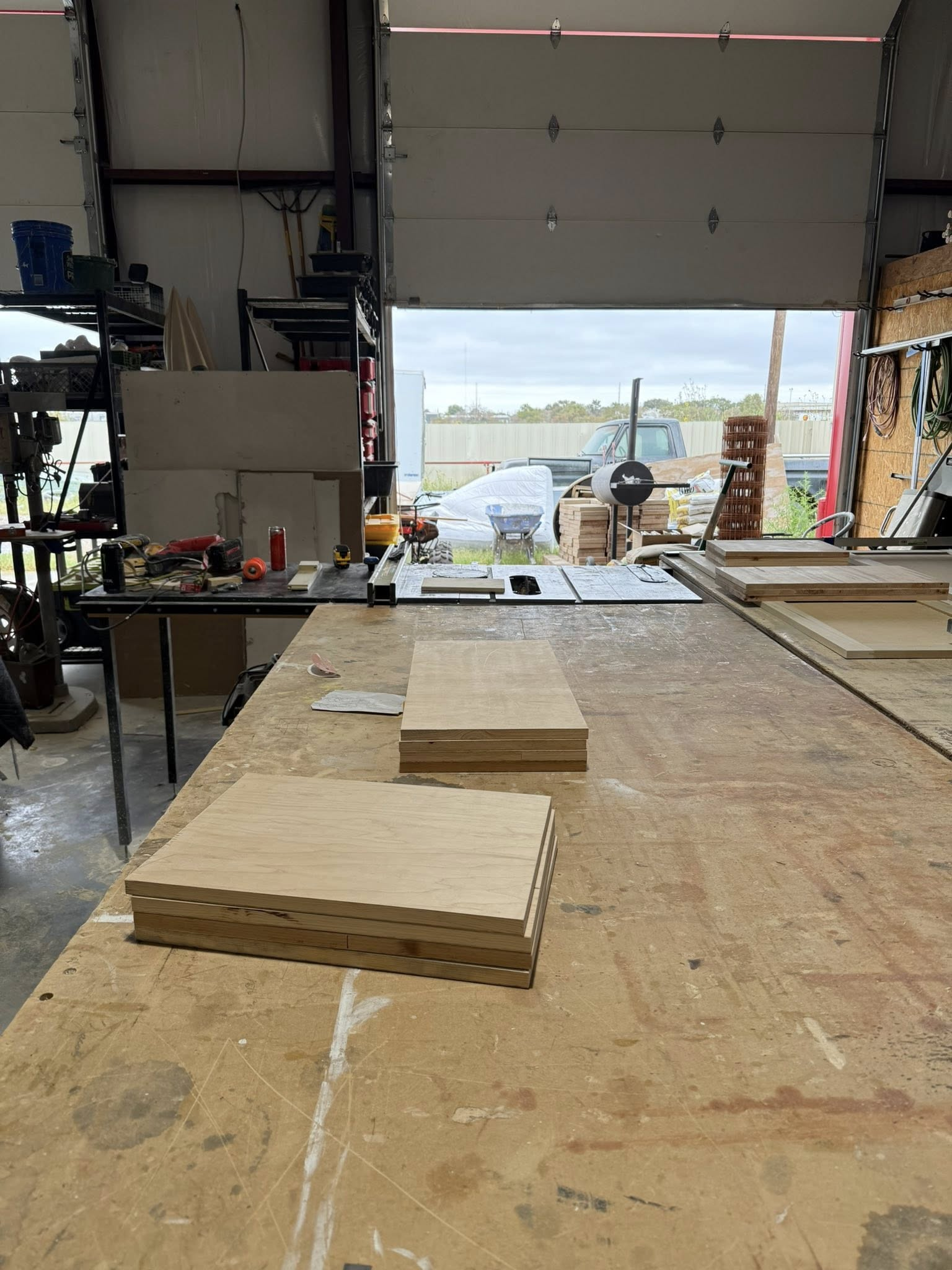 Plywood stacks on a workbench inside a shop with the door open, revealing outdoor view.