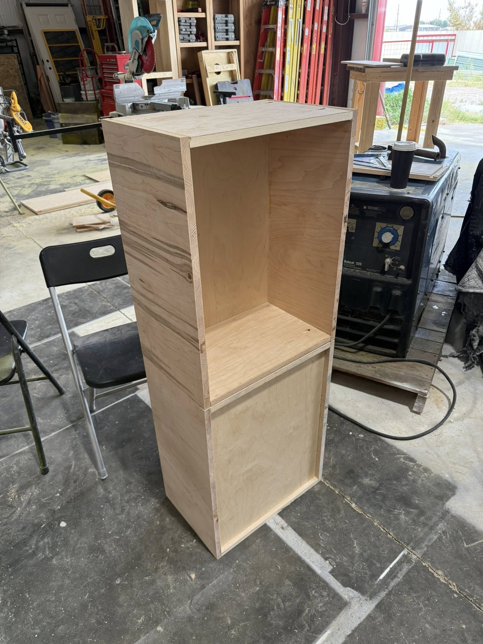 A wooden, open-faced cabinet with a shelf, standing in a workshop.
