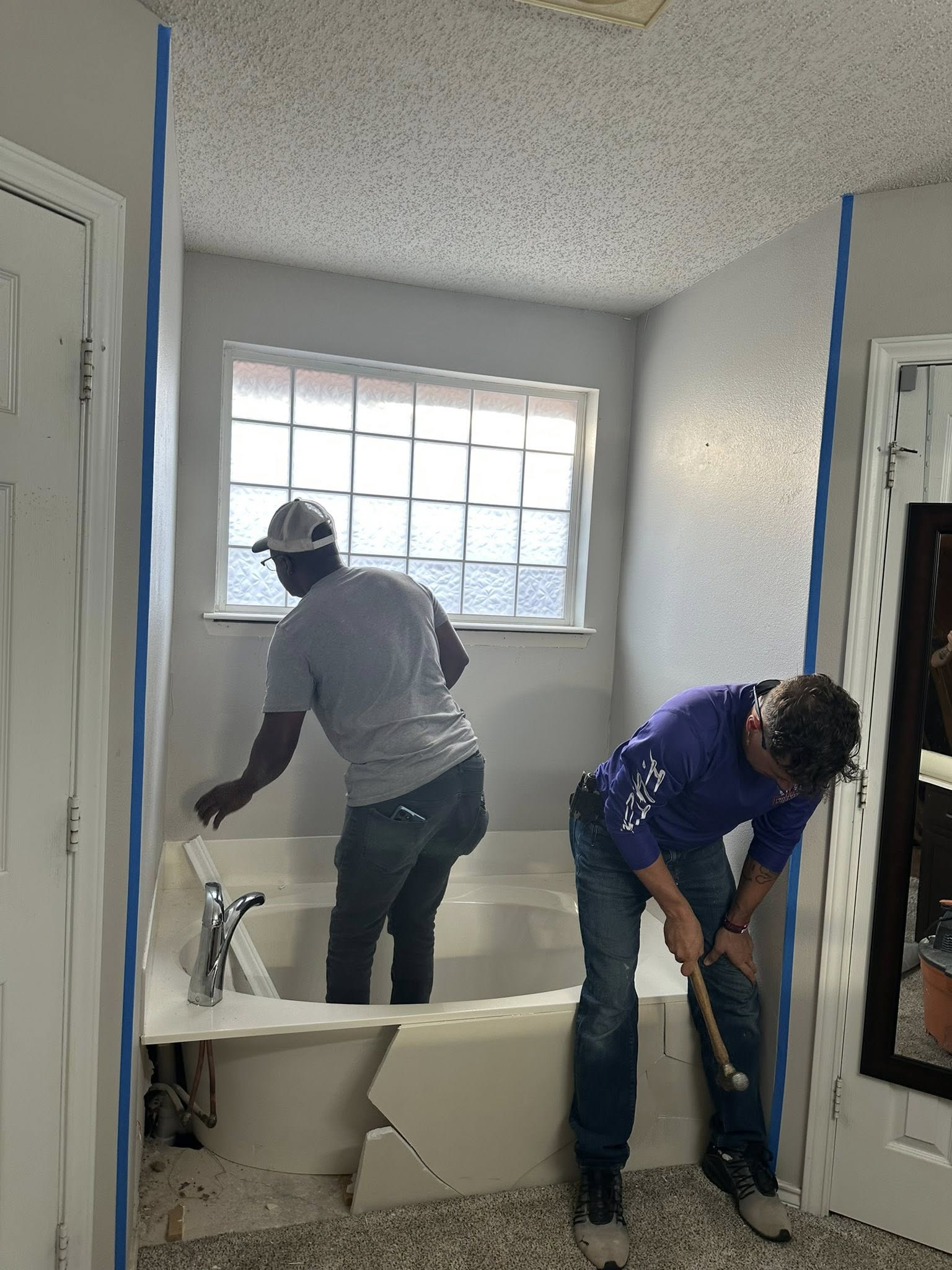 Two people renovating a bathroom; one inside the tub, the other hammering. Blue tape lines the walls.