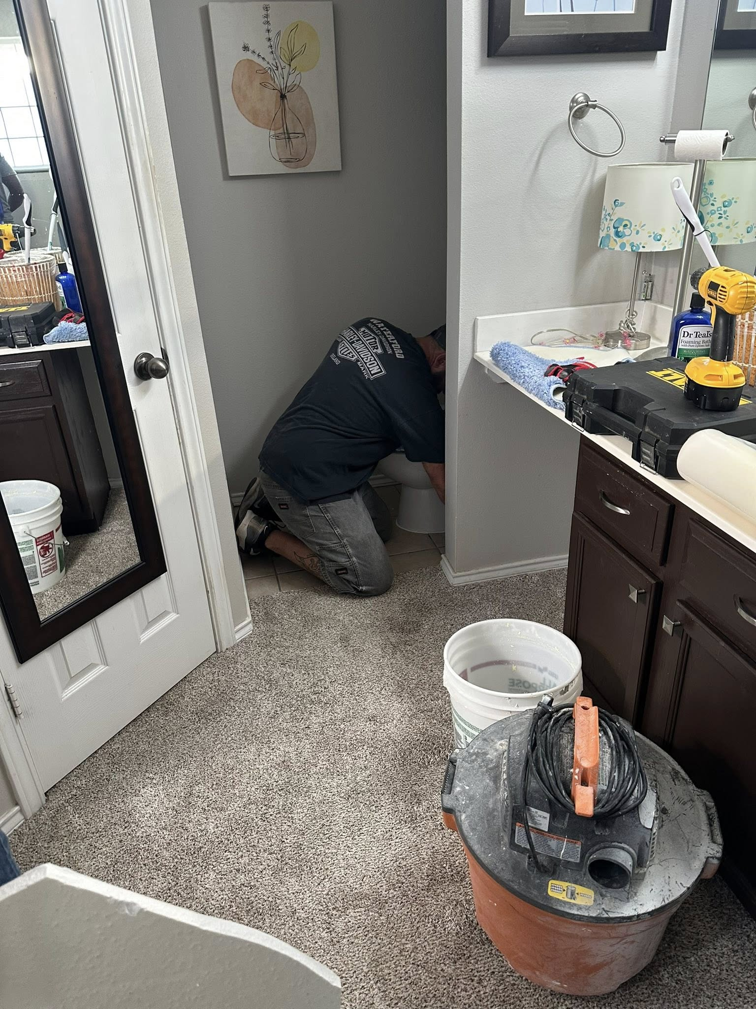 Man kneeling, working on baseboard in a bathroom. Gray walls, brown cabinets, speckled carpet.
