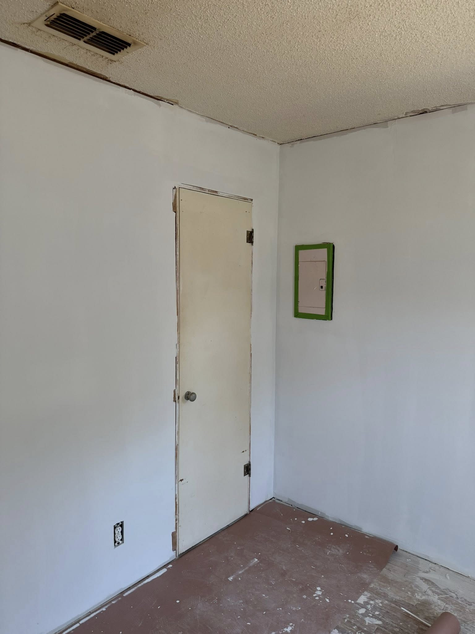 A small room with white walls, a door, and a green-taped square on the wall. Brown flooring, popcorn ceiling.