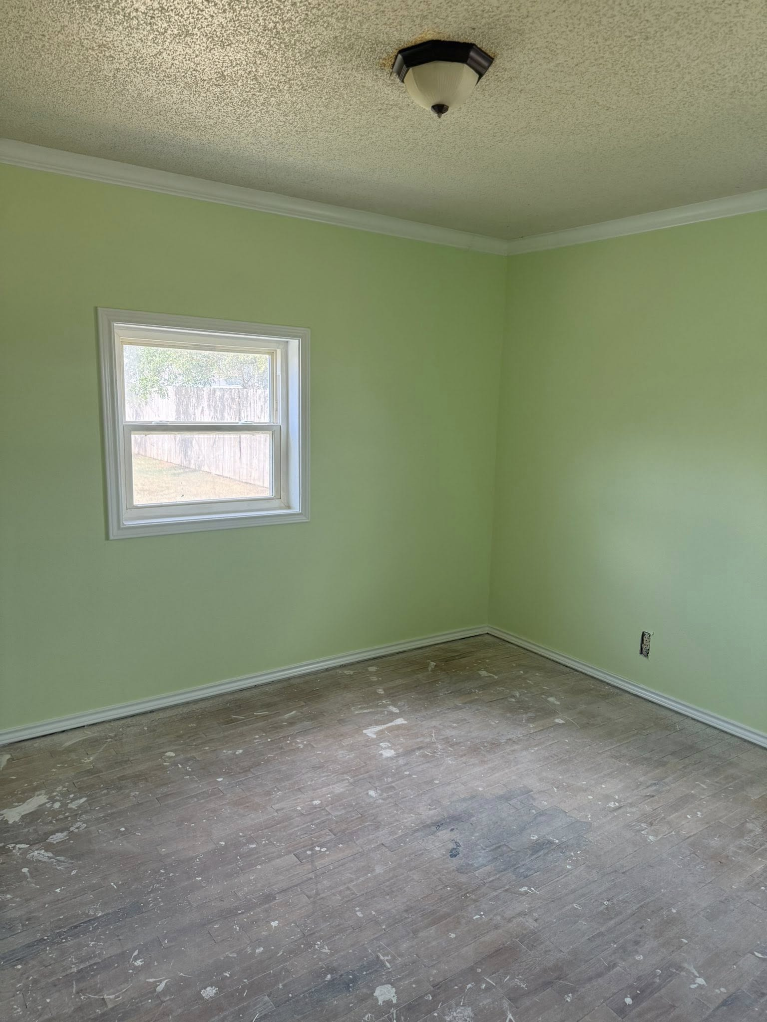 Empty room with light green walls, white trim, window, and a textured ceiling. Damaged wood-look flooring.