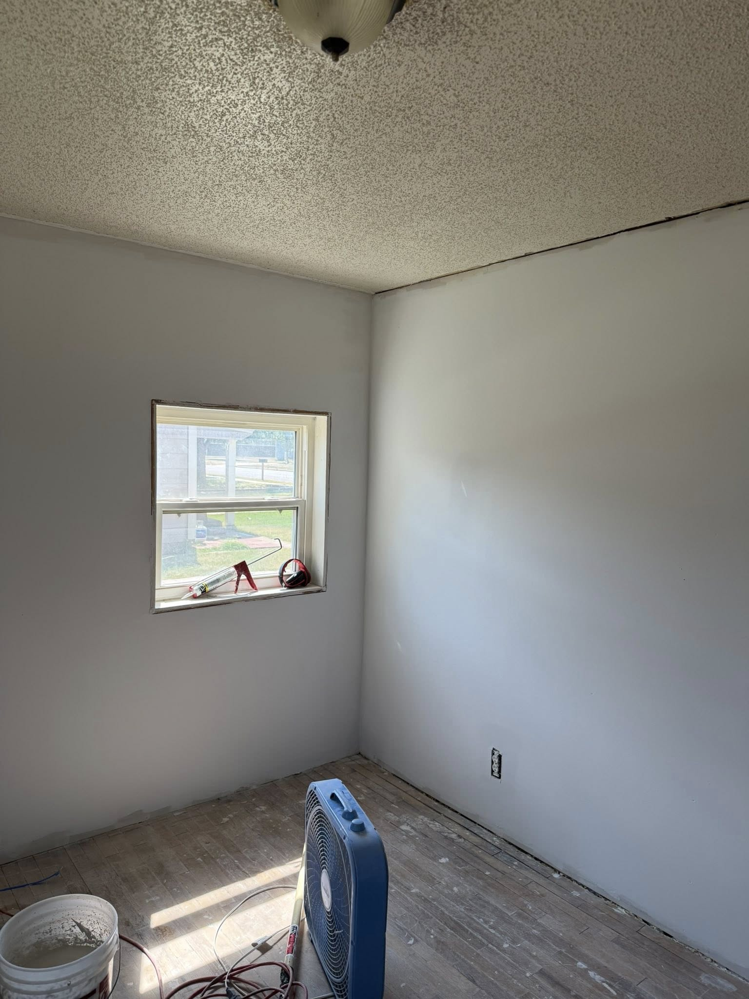 Small room with a window and a ceiling light. Gray walls, white window frame, a fan and a bucket on the floor.