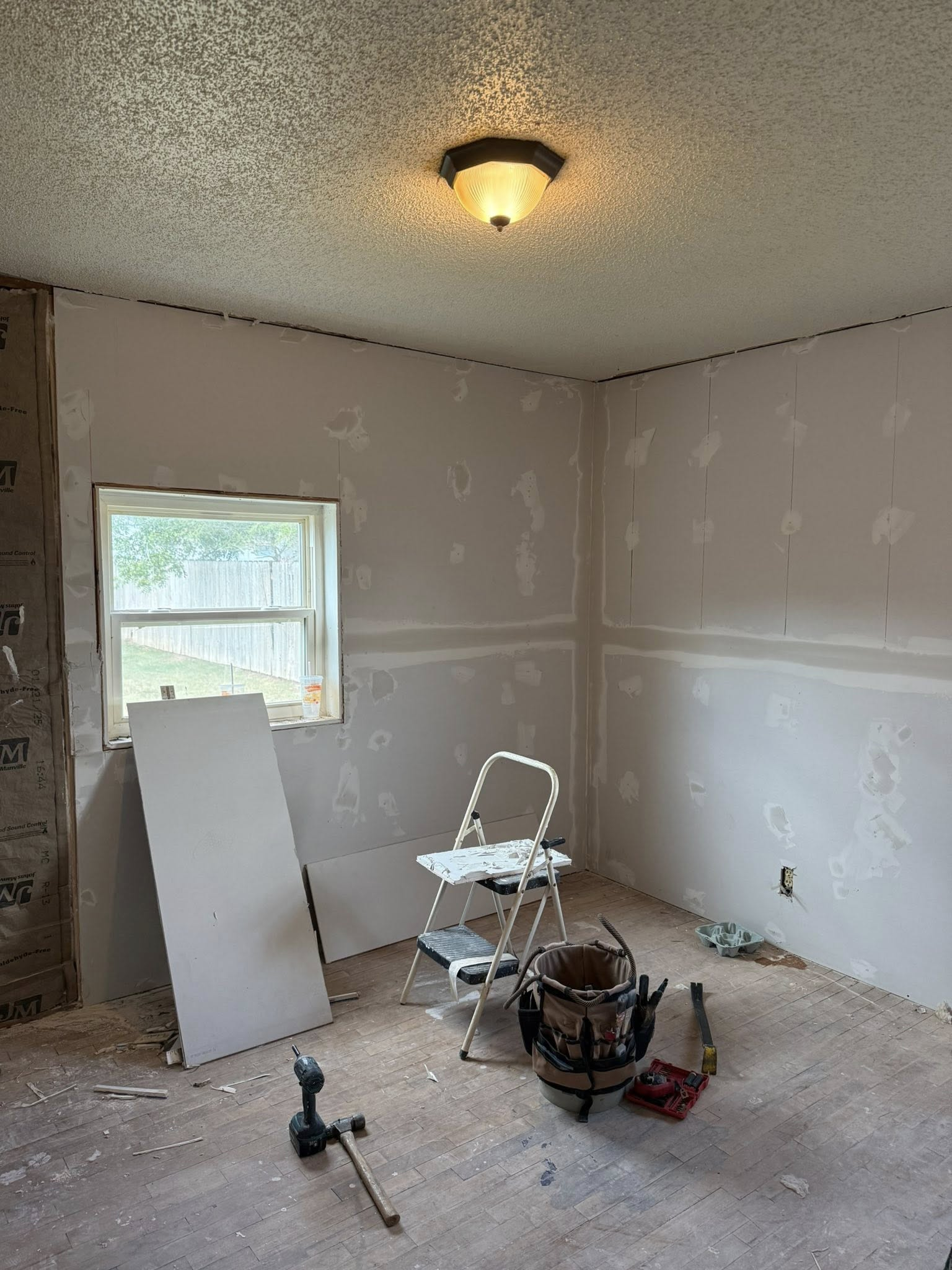 Room under renovation with drywall installed; ladder, tools, and partially covered window.