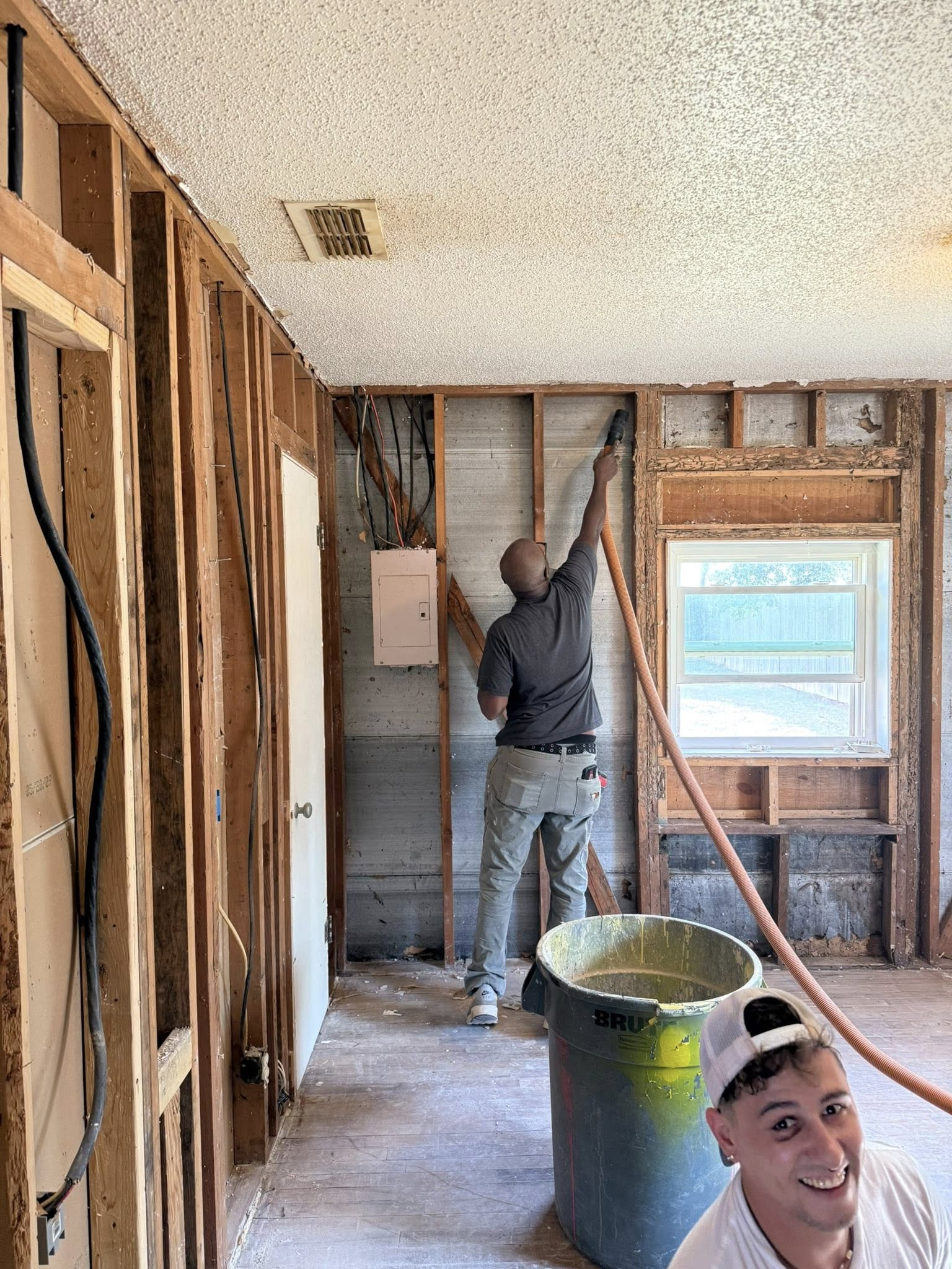 Man spraying insulation in a room with exposed wooden studs, wearing a hat, and a person in the lower right corner.