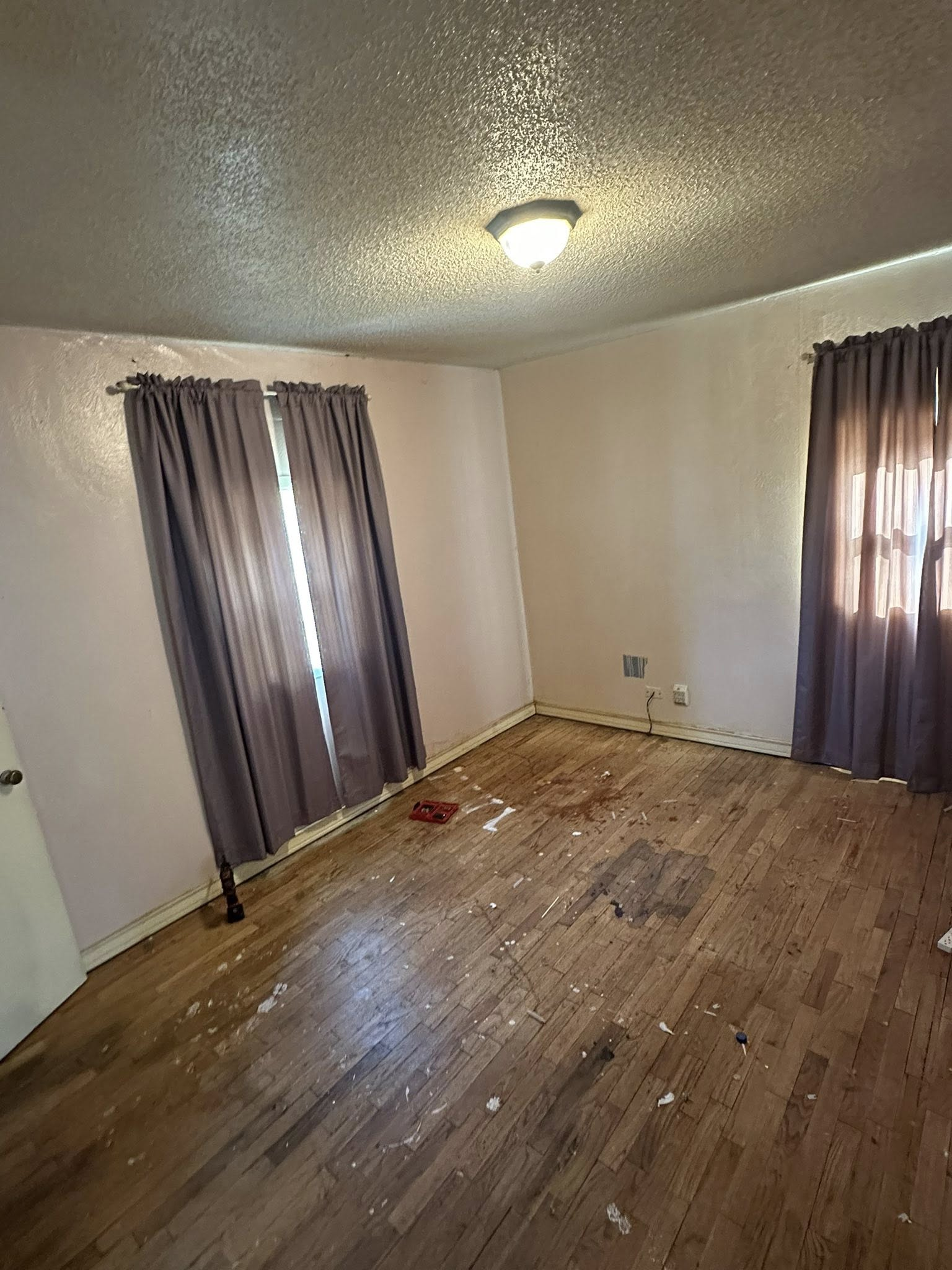 Empty room with wooden floors, two windows with purple curtains, and a ceiling light.