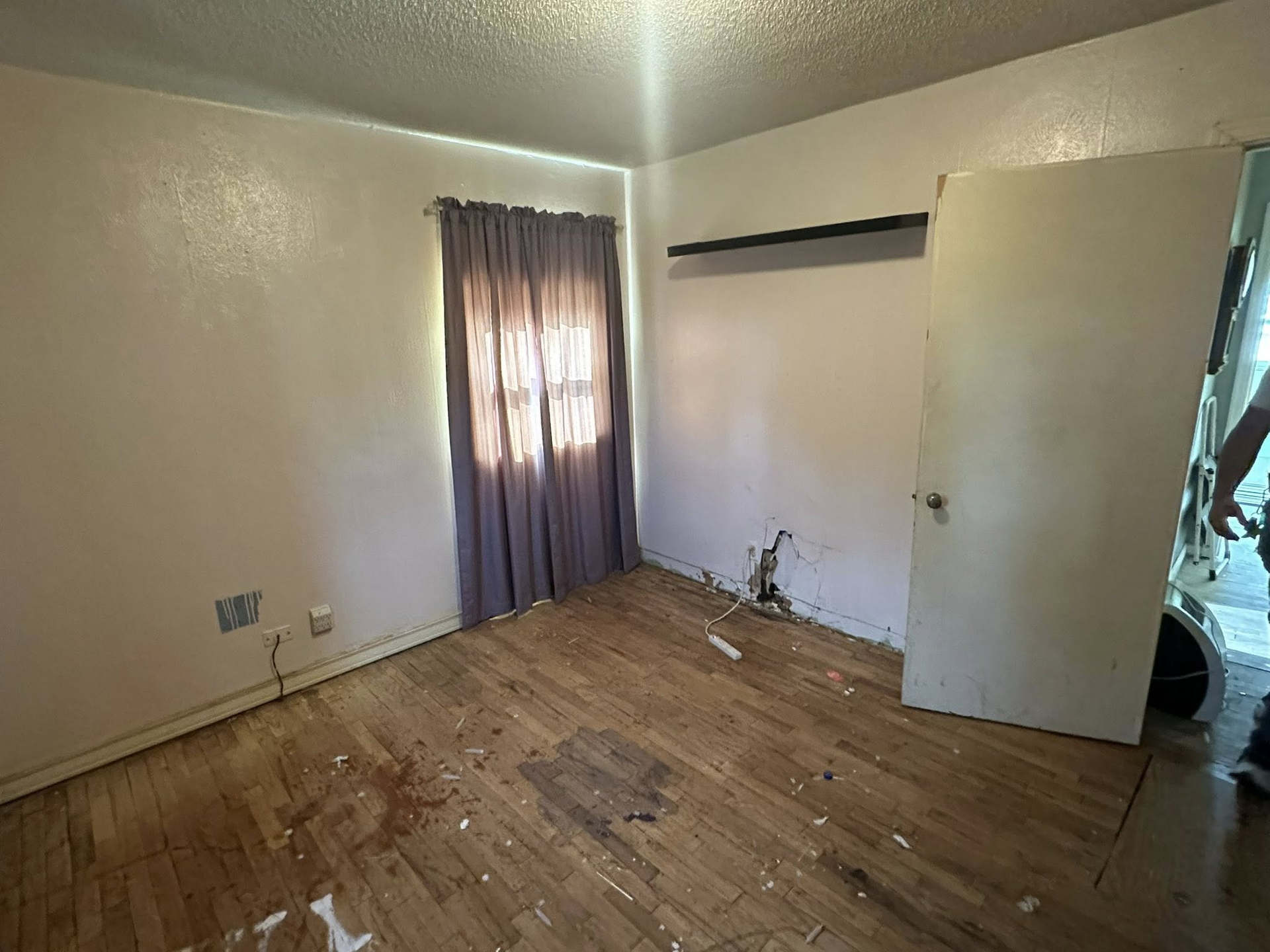 Empty room with worn wooden floor, off-white walls, window with purple curtains, open white door.