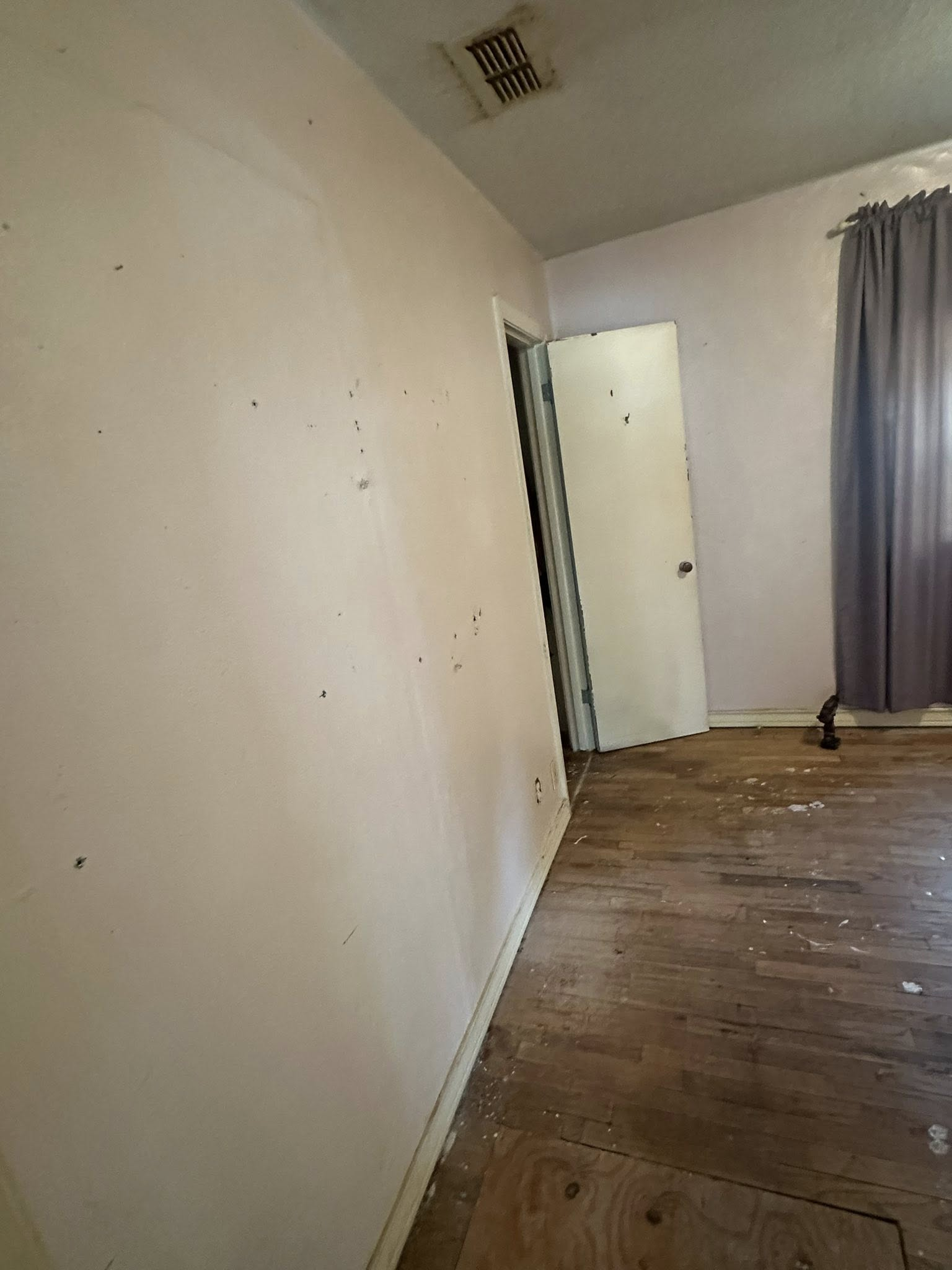 A room with peeling floor, white walls, and a closed door. Grey curtain visible.
