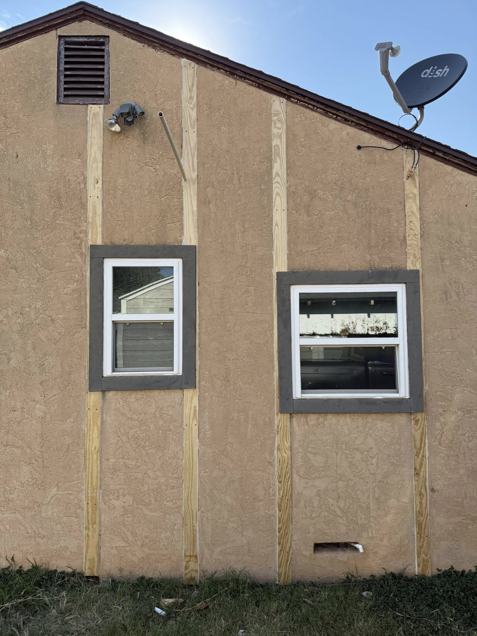 Tan stucco house side with two windows, wood trim, satellite dish, and ventilation vent.