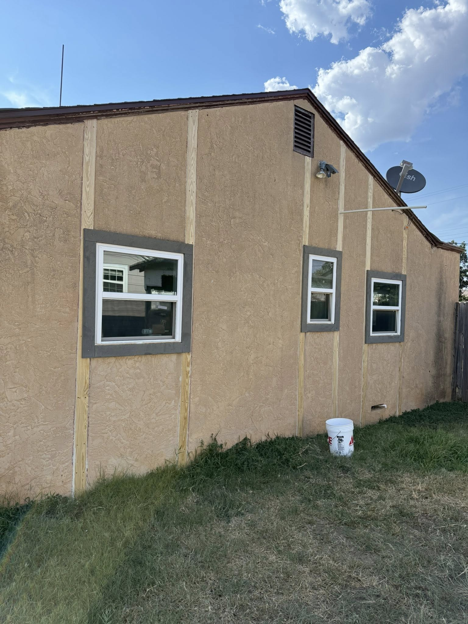 Side view of a house with beige stucco, white-framed windows, and tan accents; bucket sits below.