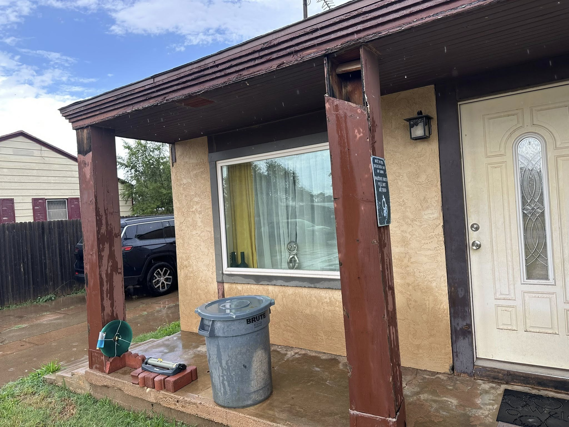 Exterior of a house in rain; a porch with damaged wood, a trash can, and a partially visible car.