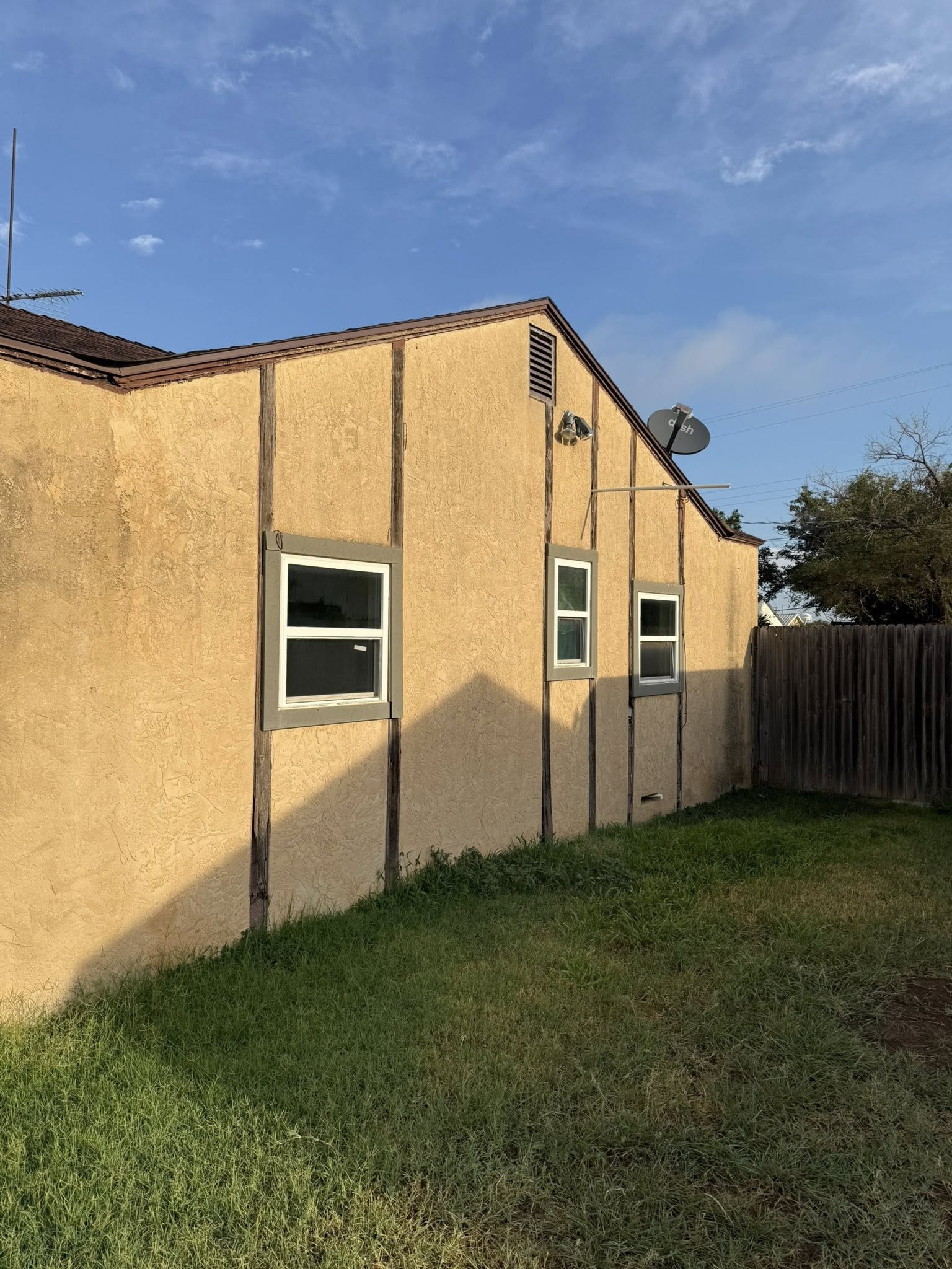 Side of a stucco house with brown trim, three windows, and a small grassy lawn under a blue sky.
