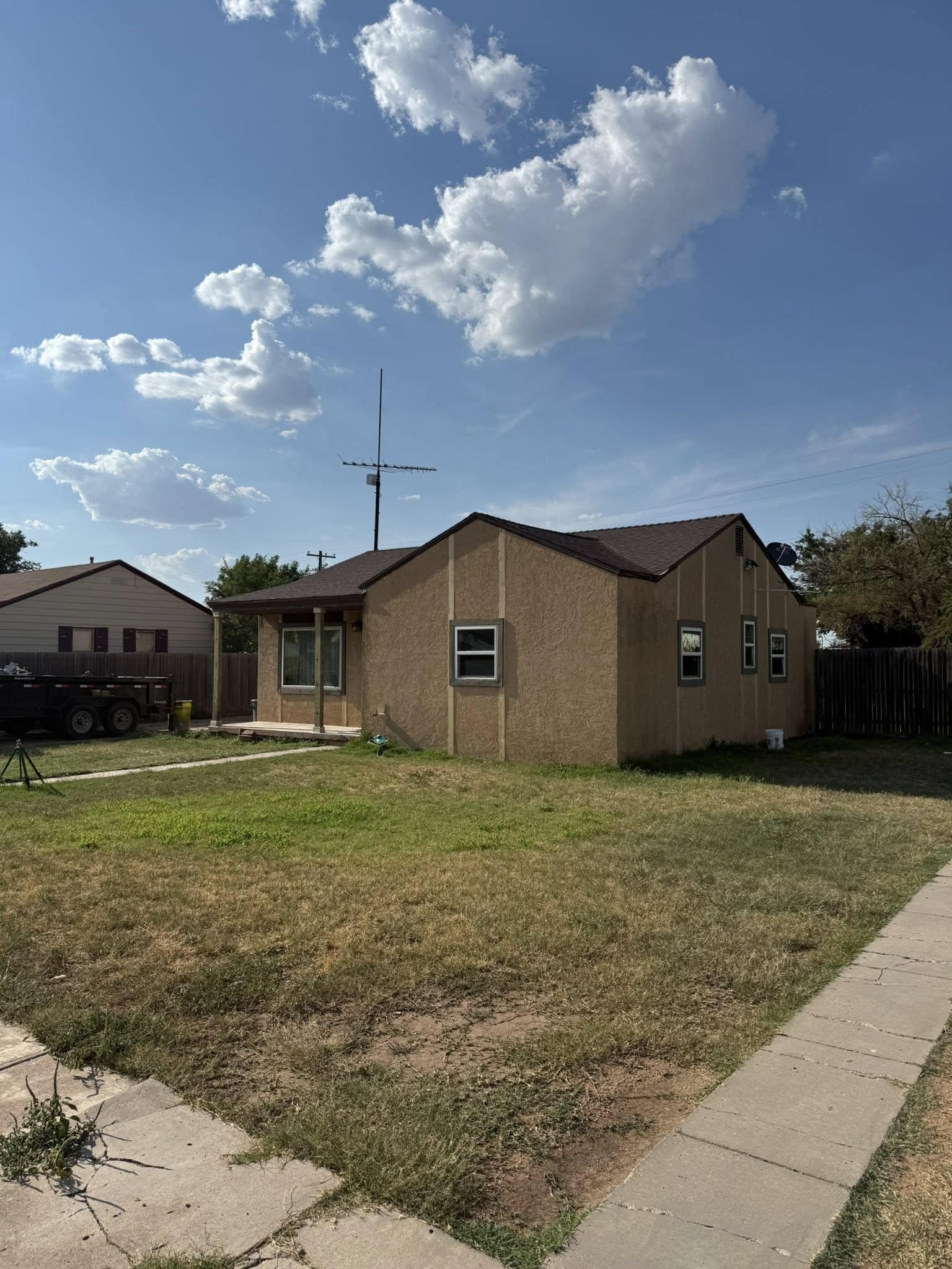 A one-story tan house with brown roof and a grassy lawn under a blue sky with puffy clouds.