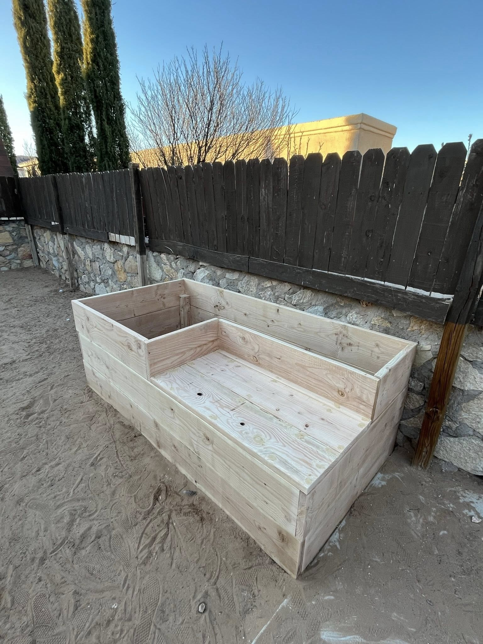 Wooden raised garden bed in an outdoor setting, with a dark fence and blue sky background.