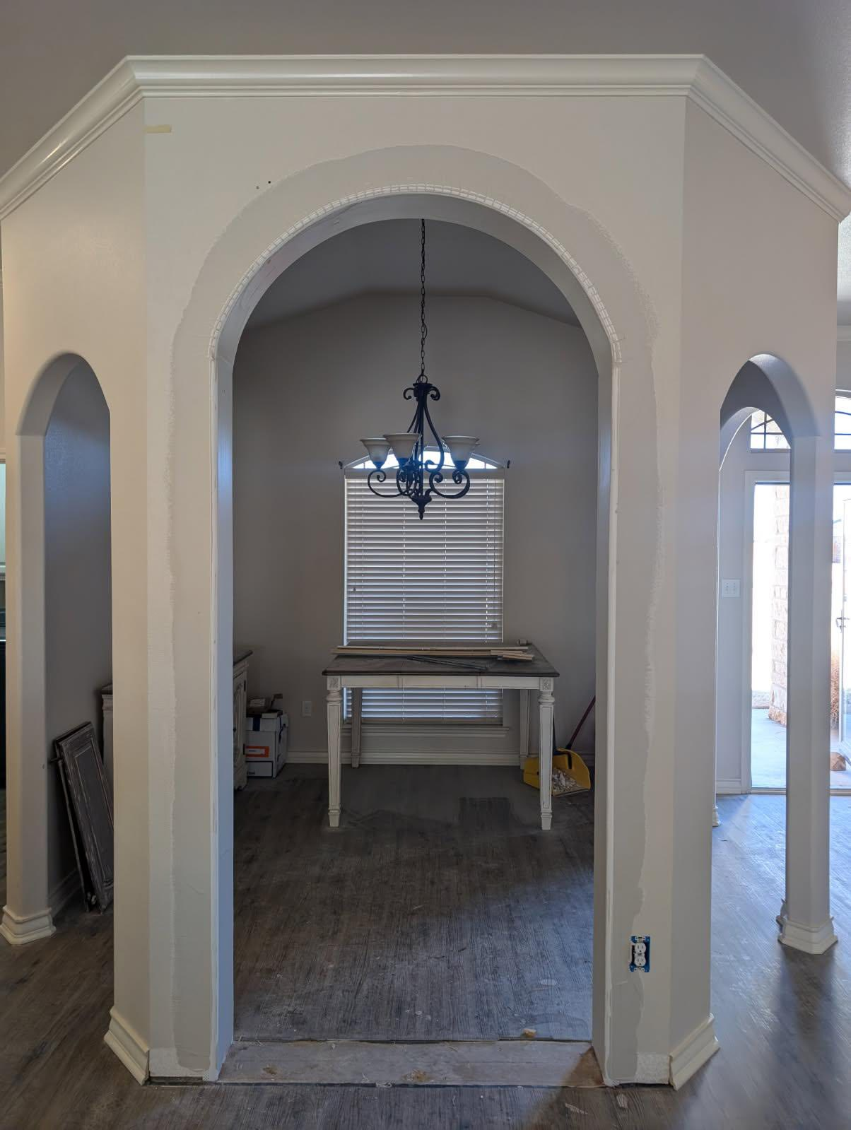 White arched doorways leading into a dining room with a chandelier, table, and window with blinds.