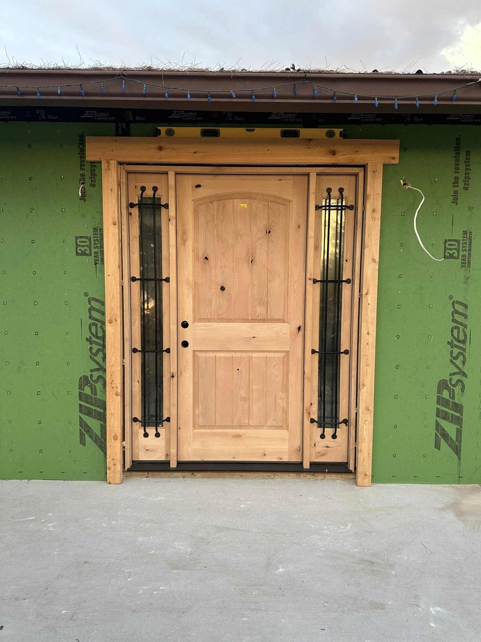 Wooden door with sidelights, black ironwork, rustic trim, set against green siding.