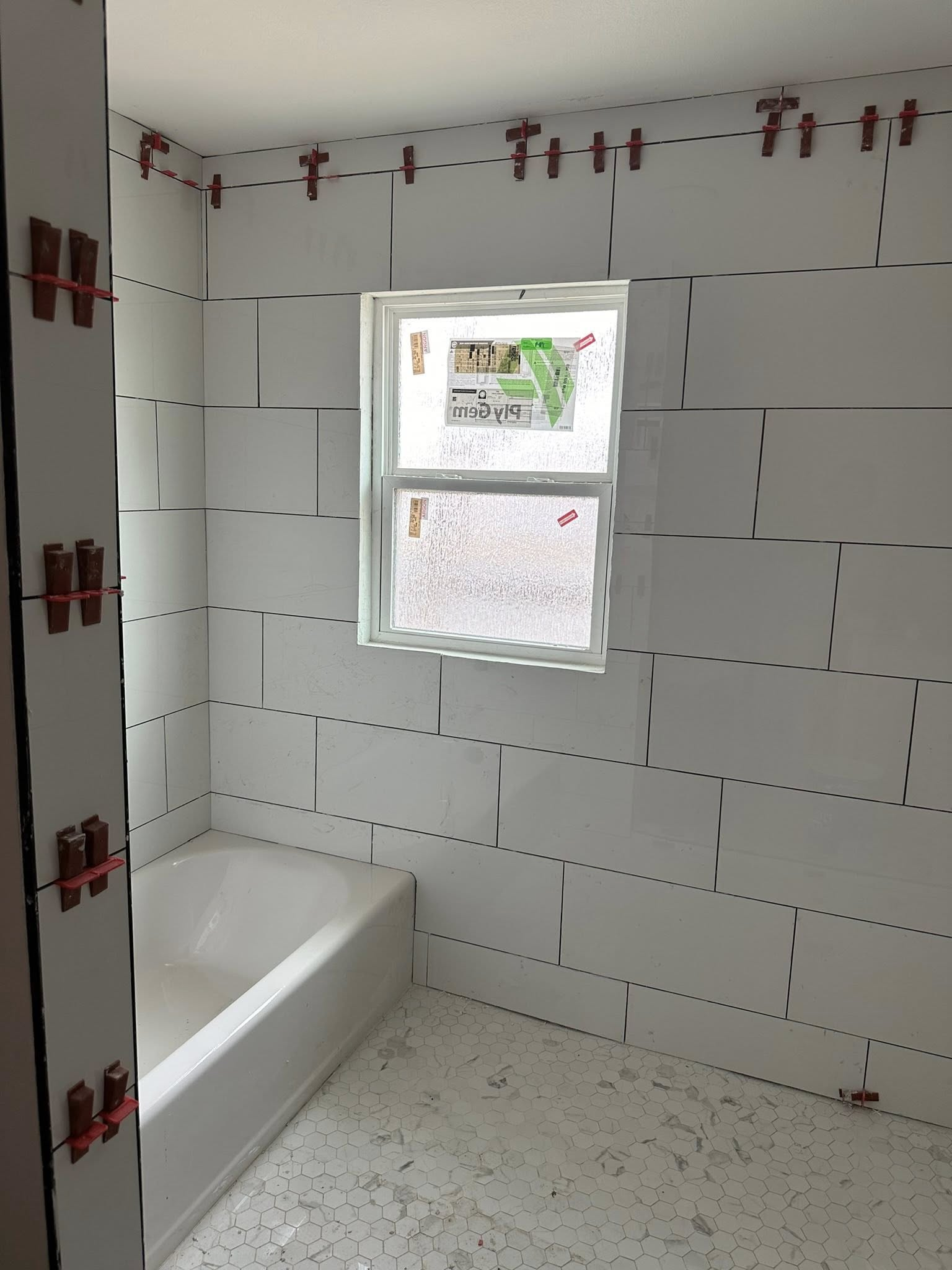 Bathroom in progress with white tiles and tub, a window, and tile leveling clips.
