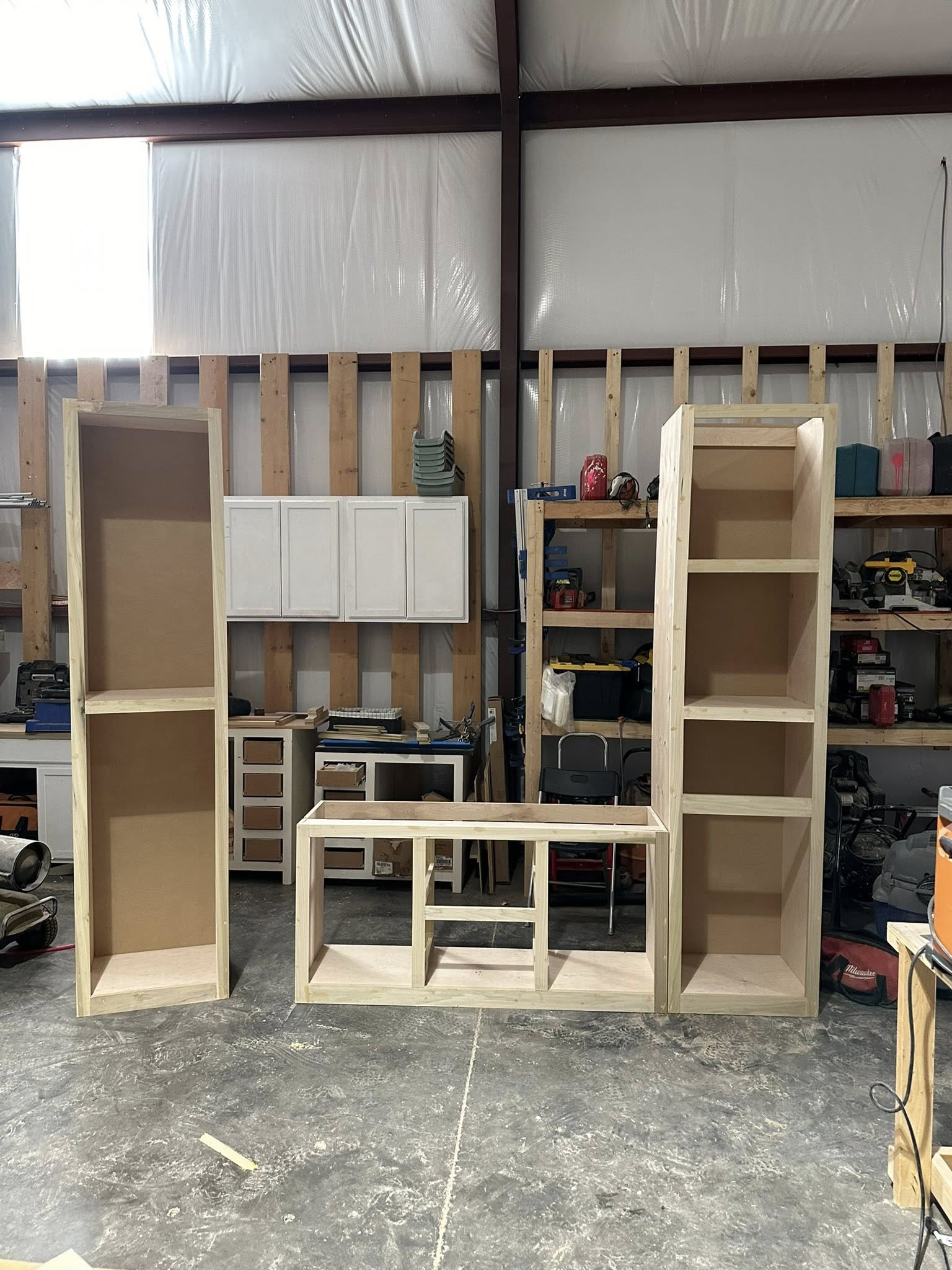 Unfinished wooden cabinets in a workshop, including two towers and a low cabinet, with white cabinets in the background.