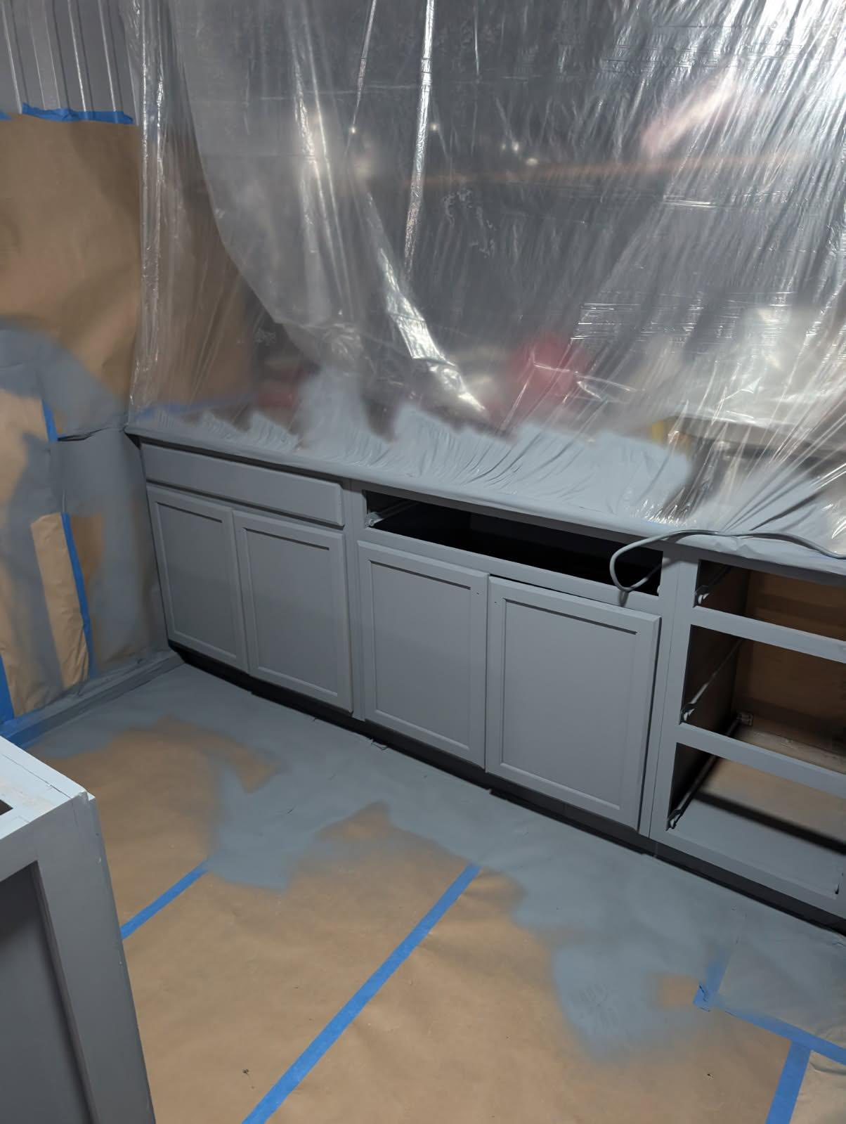 Kitchen cabinets partially painted gray, covered with plastic sheeting, in a room prepped for painting.