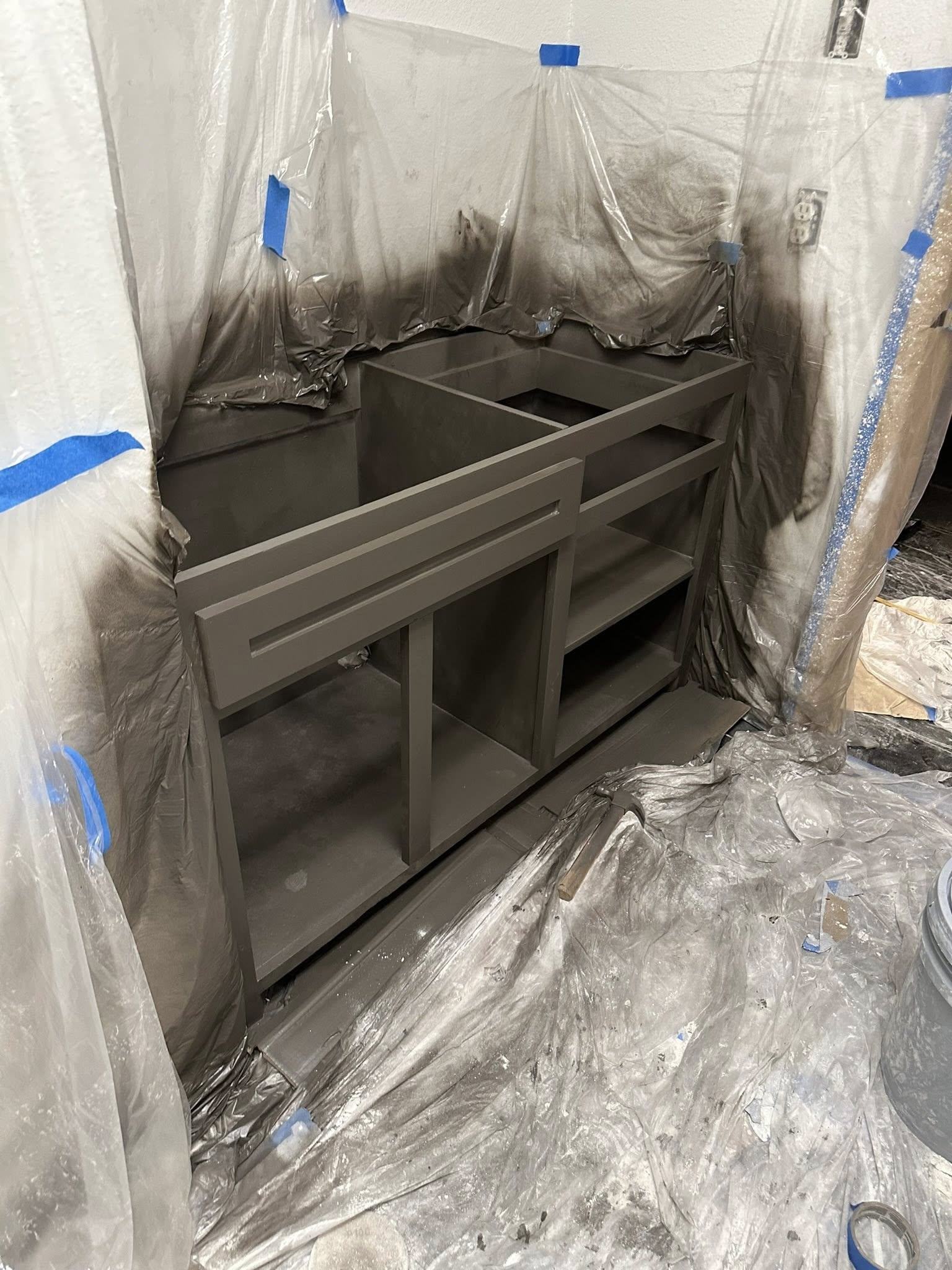 Gray-painted cabinets covered in plastic sheeting, surrounded by paint splatters; room under renovation.