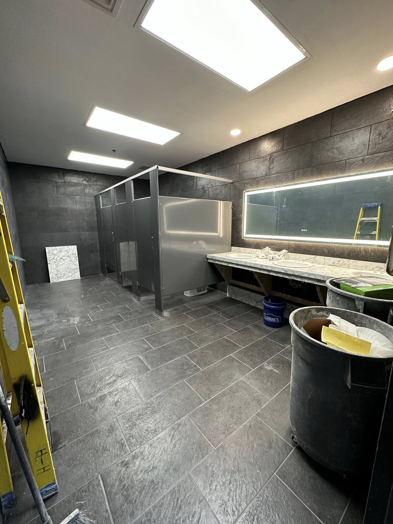 Bathroom under construction with stalls, sinks, gray tile, and dark gray walls.