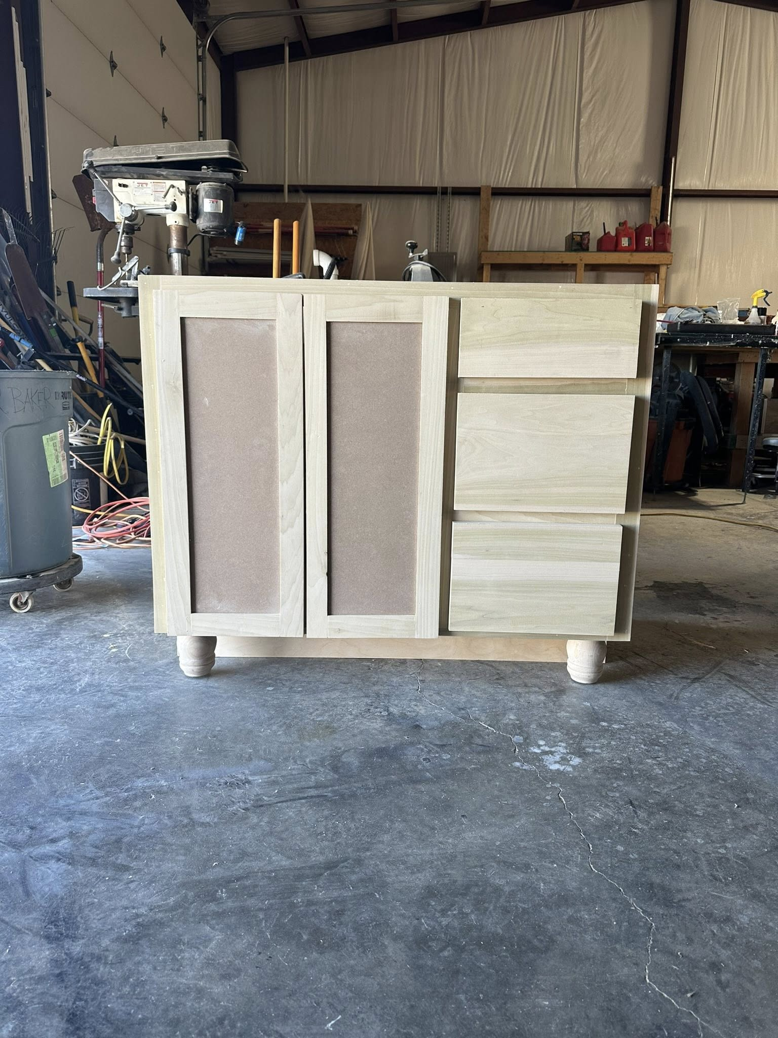 Unfinished wooden bathroom vanity cabinet with two doors, three drawers, and round feet in a workshop.