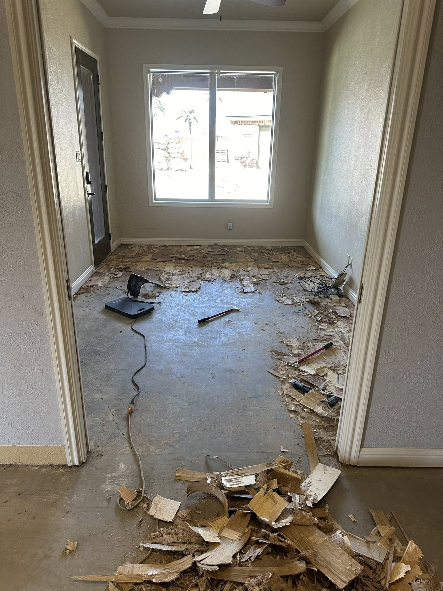 Room with debris on the floor, door on left, window on back wall, and concrete floor visible.