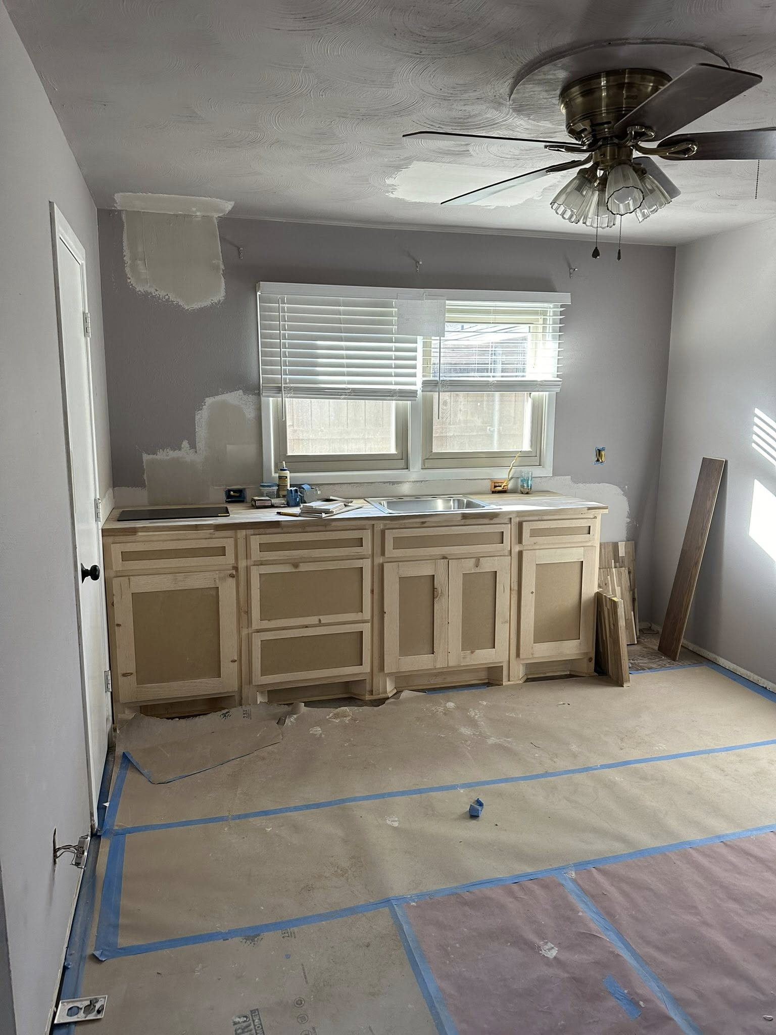 Room renovation in progress: unfinished cabinets, window, ceiling fan, protective floor covering, light gray walls.