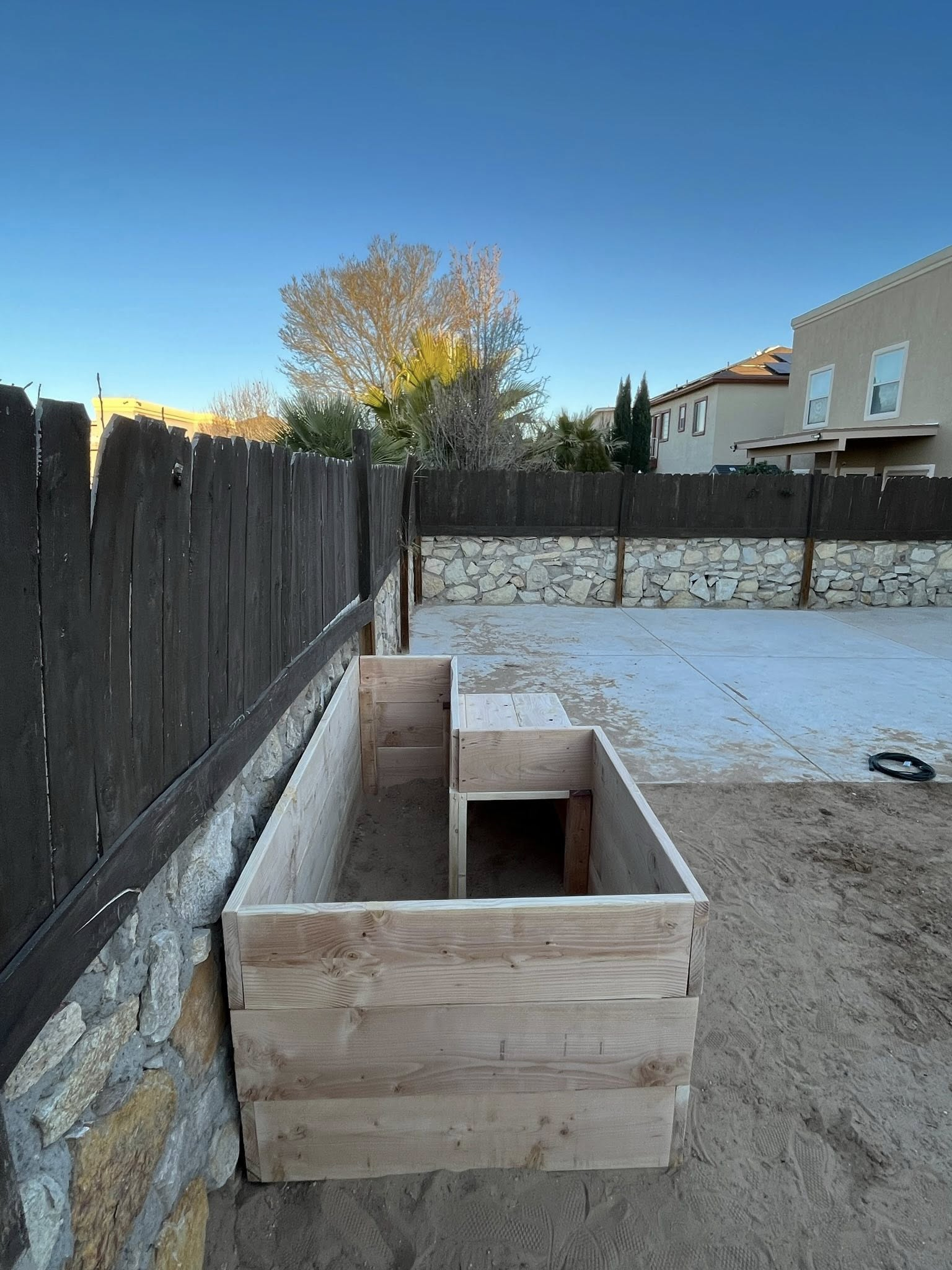 Wooden box planter next to a brown fence and stone wall in a backyard on a sunny day.