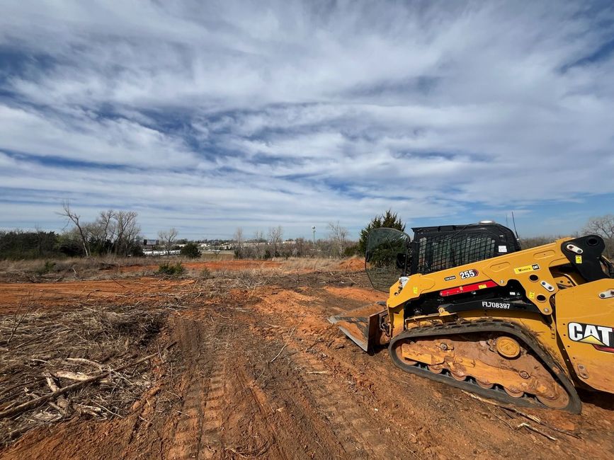 A yellow Caterpillar skid steer loader sits on a cleared, red-dirt construction site under a bright, cloudy sky.