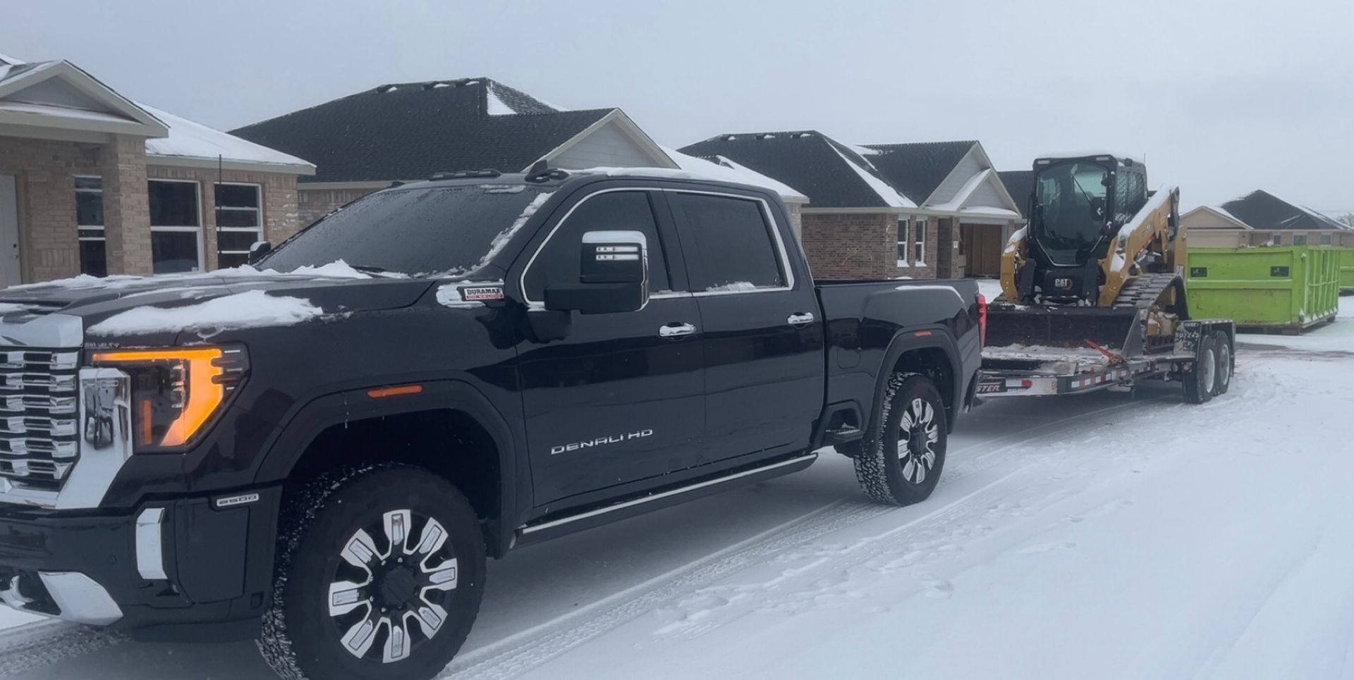 A black pickup truck parked in snow, towing a flatbed trailer with a yellow skid steer and a green dumpster.