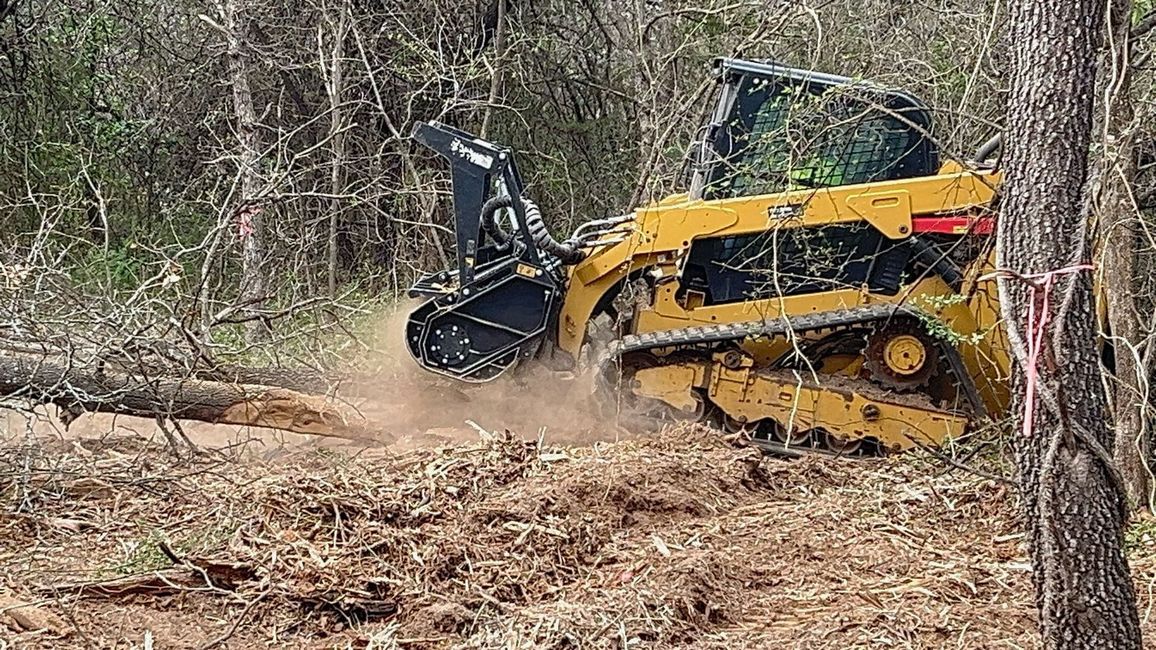 A yellow track loader mulcher shreds a fallen log in a wooded area, kicking up debris.