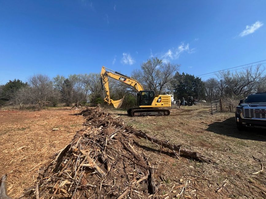A yellow Caterpillar excavator sits on a clearing in a field, near a large pile of brush and a pickup truck.