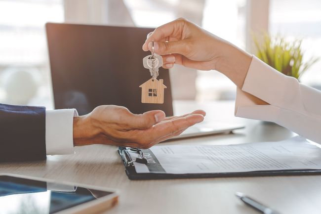 Person handing over house keys to another person. Desk with laptop and papers.
