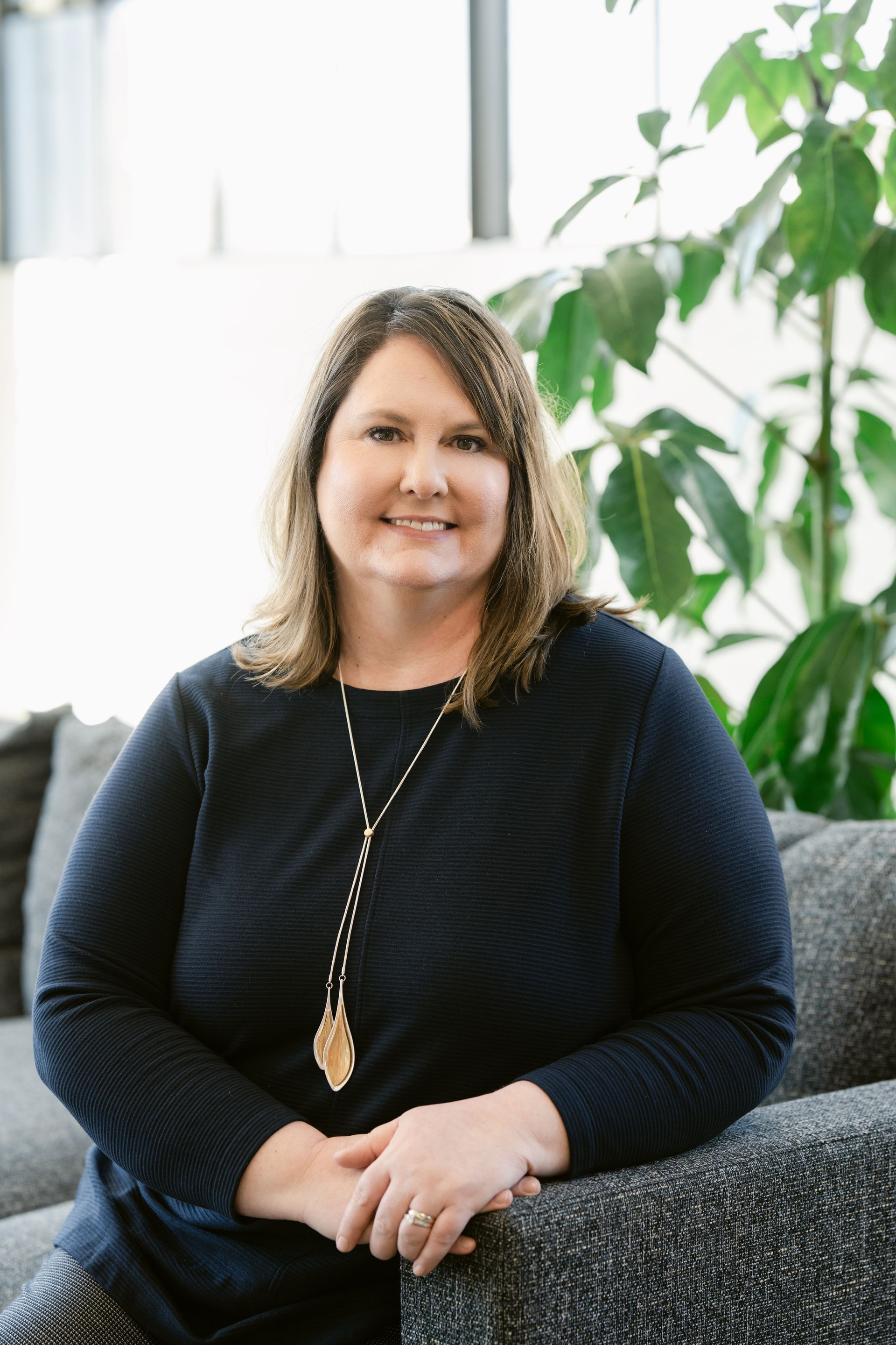 Melissa Morris with shoulder-length brown hair, smiling, wearing a navy sweater and necklace, sitting on a gray couch in front of plants.