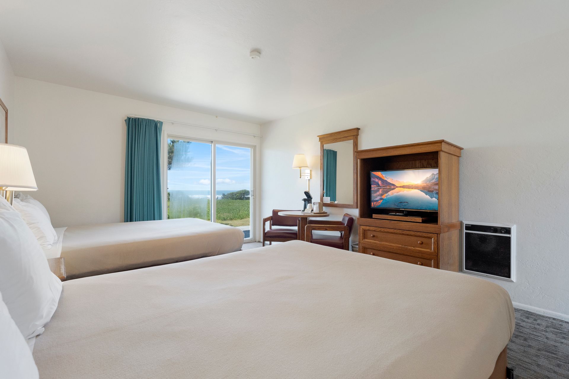Hotel room with two beds, a TV, desk, and a view of the ocean through a sliding glass door.