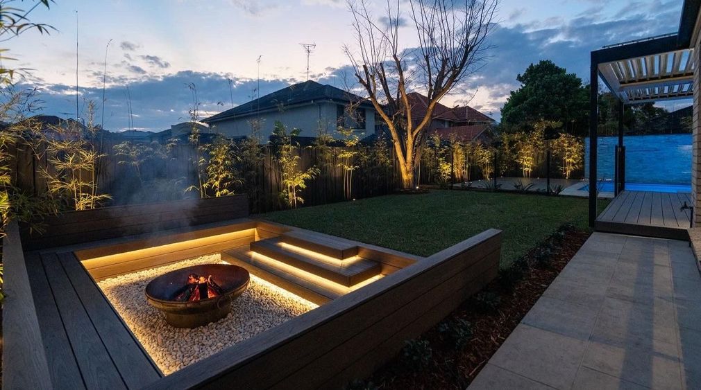 Modern backyard patio at dusk with a sunken fire pit, illuminated steps, and garden seating area