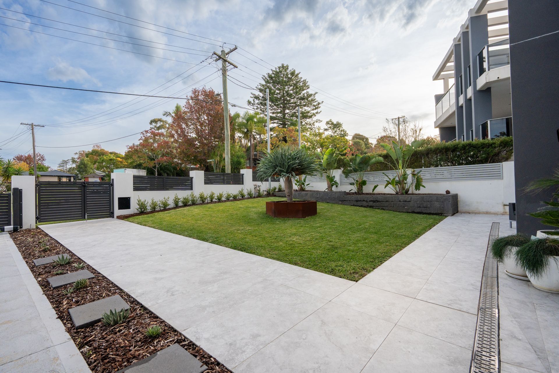 Driveway and lawn with modern landscaping; grey pavers, black gate, green grass, and low shrubs.