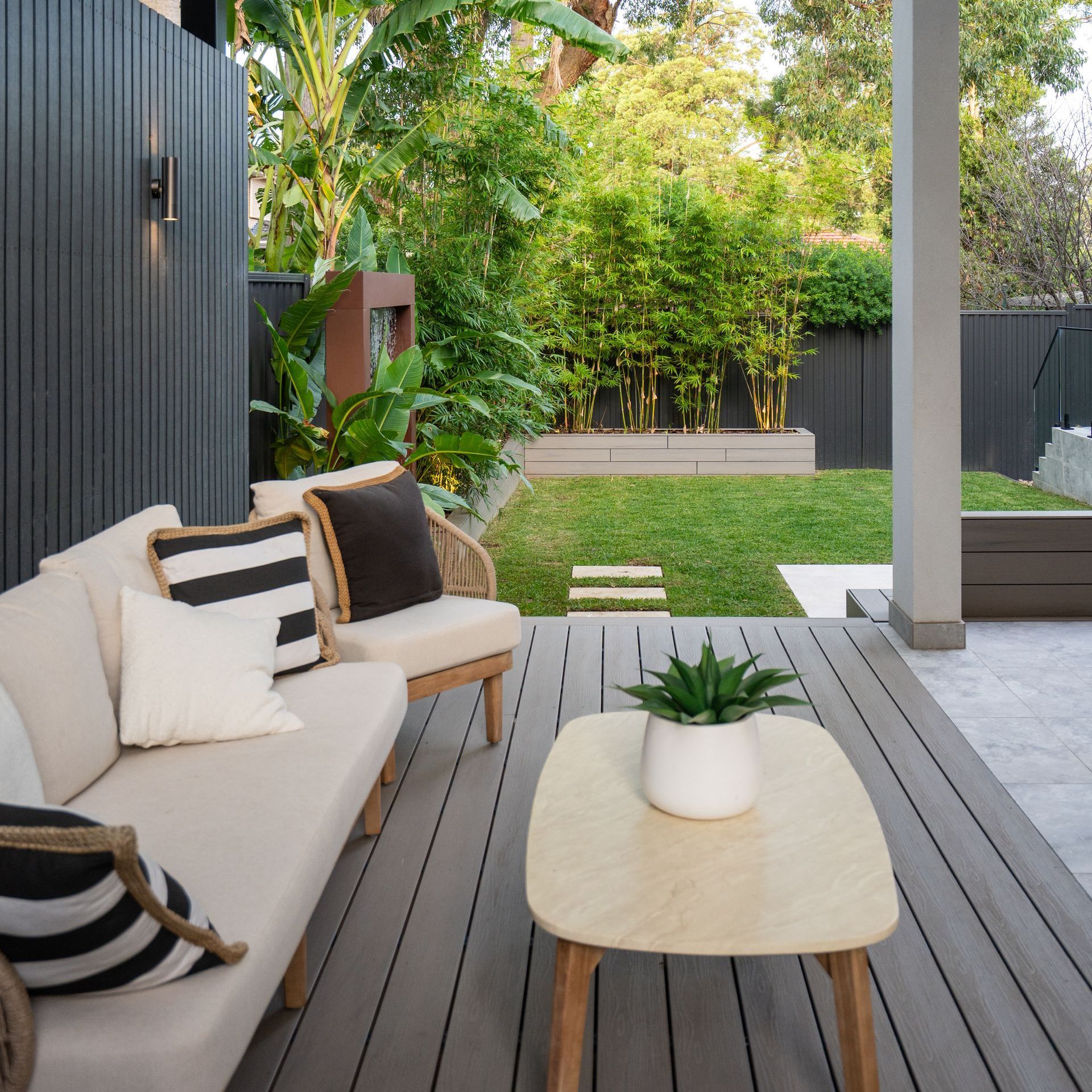 Outdoor patio with neutral-toned furniture, wooden deck, and manicured backyard with lush greenery.